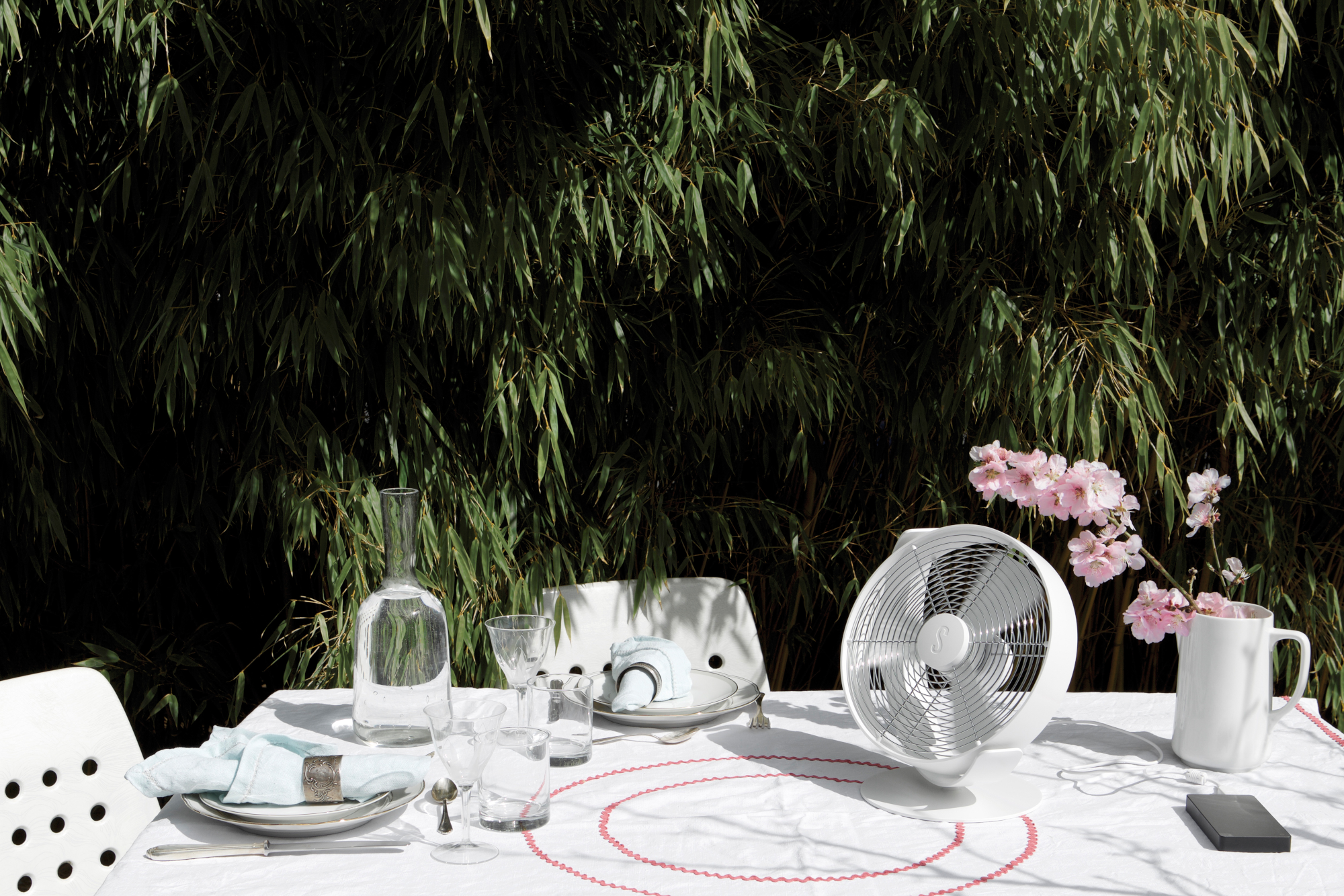 Un tavolo apparecchiato, con un ventilatore bianco e fiori. Bambù sullo sfondo.