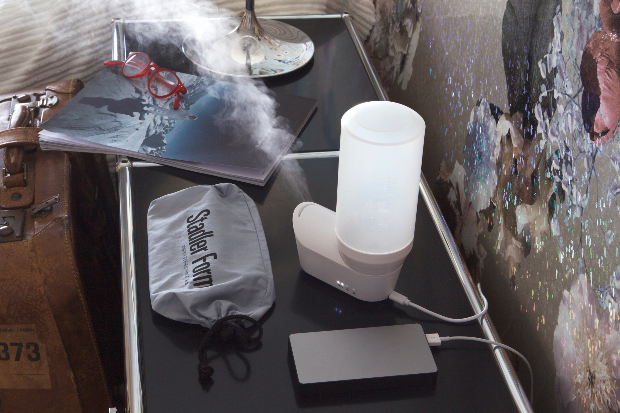 Aromadiffusor mit Dampf auf einem schwarzen Tisch. Gläser, ein Notizbuch und eine Tasche sind auch darauf.