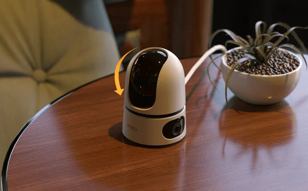 Kamera bezpieczeństwa na stole z obiektywem obrotowym, obok rośliny doniczkowej.