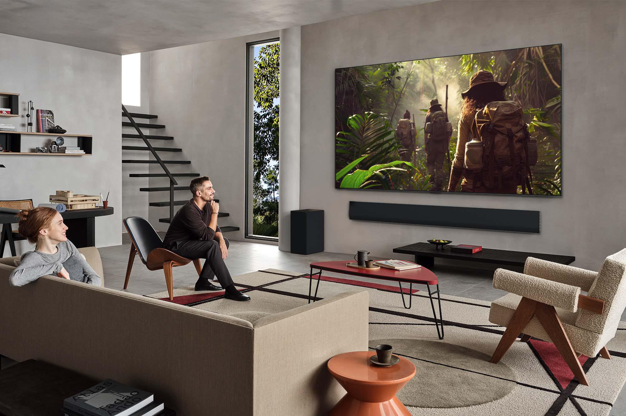 Wohnzimmer mit TV, der eine Dschungel-Szene zeigt; Paar schaut zu. Schwarze Soundbar und Subwoofer.