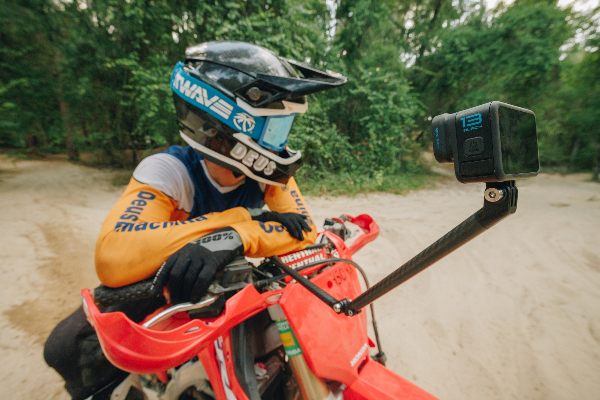 Eine Person mit Helm und GoPro-Kamera an einem Motorrad. Der Hintergrund zeigt Bäume und Sand.