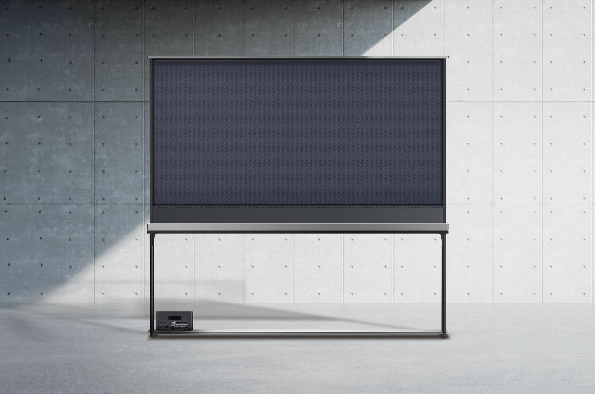 Eine Projektionsleinwand auf einem Ständer vor einer Betonwand. Unter der Leinwand befindet sich eine Steuerbox auf dem Boden.