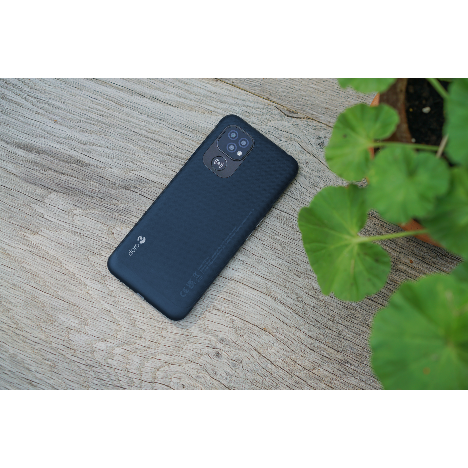 A dark blue Doro smartphone lies on a wooden surface. Green plant leaves and a pot are on the right side of the image.