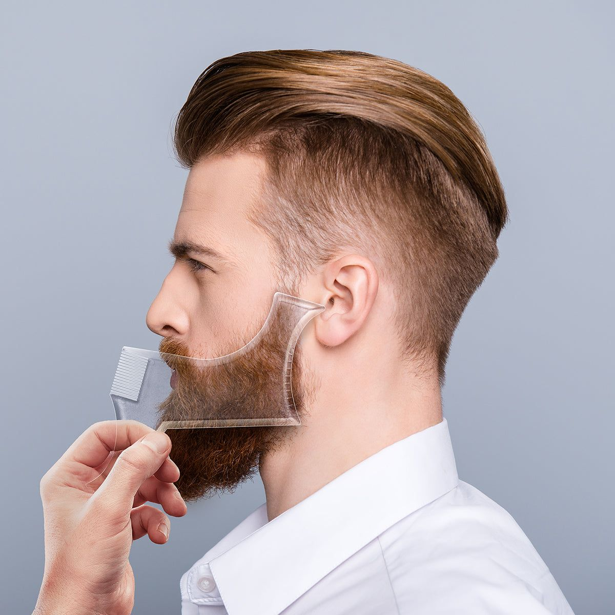 Hombre con barba, usando herramienta para dar forma a la barba, fondo gris.