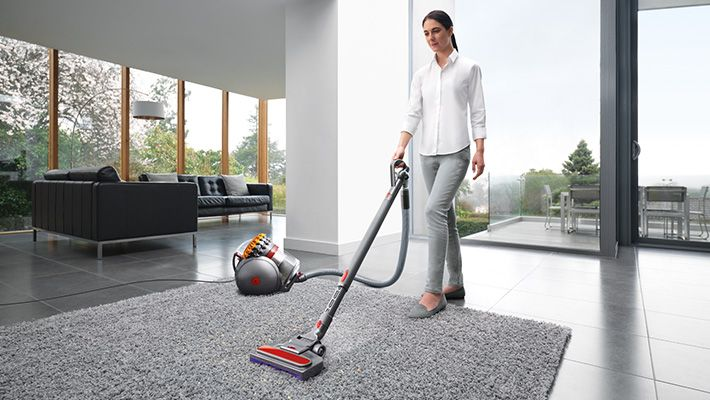 Kobieta odkurzająca szary dywan srebrno-czerwonym odkurzaczem. Salon z oknem w tle.