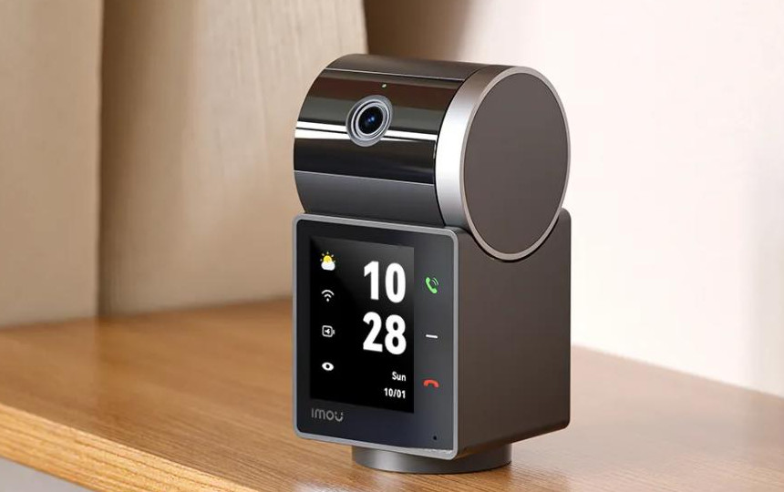 Czarno-srebrna kamera bezpieczeństwa z ekranem wyświetlającym godzinę i pogodę na drewnianej półce.