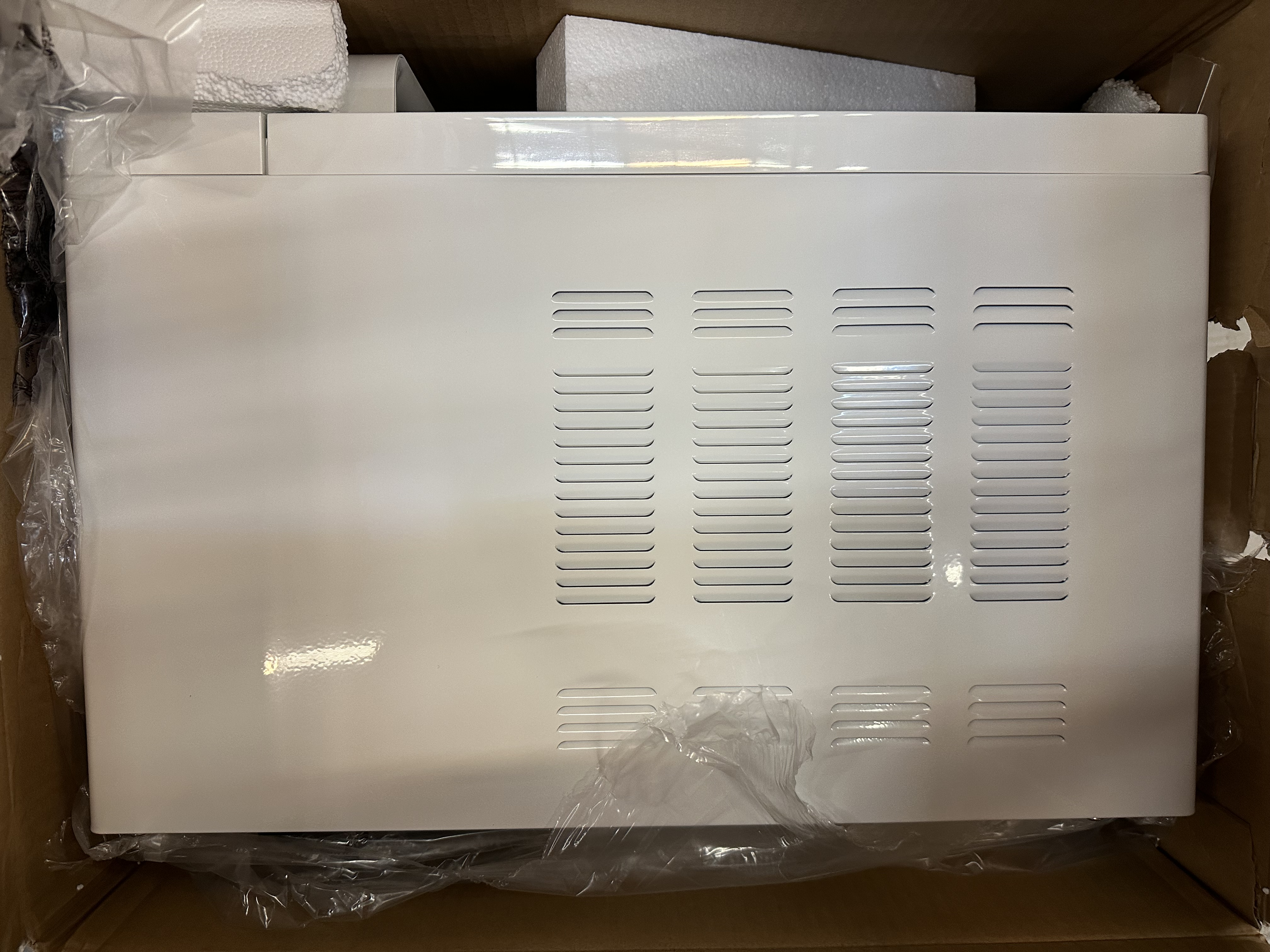 Białe urządzenie w pudełku z opakowaniem styropianowym. Otwory wentylacyjne z tyłu.