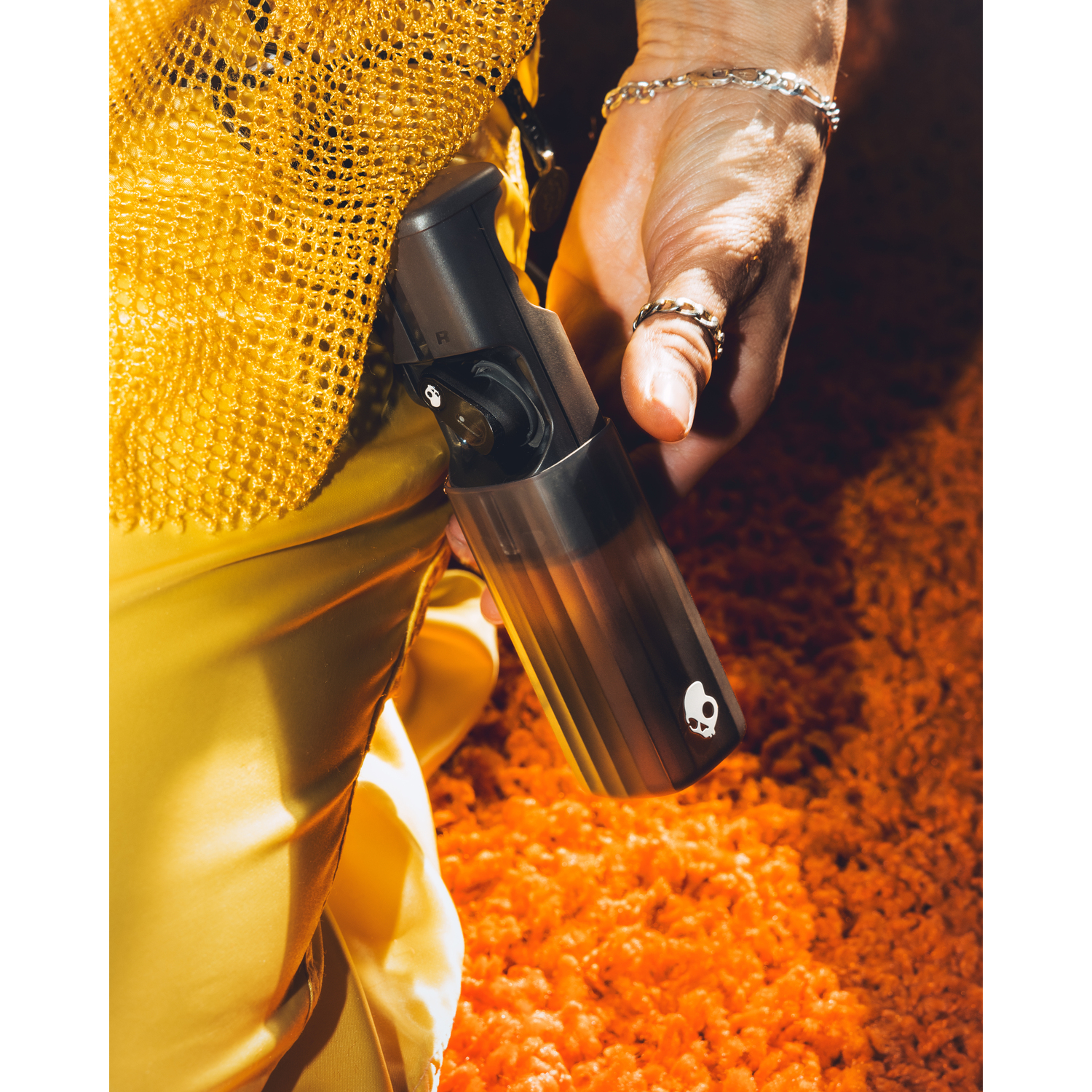 Person hält schwarzen Ohrhörer-Etui, orangefarbener Hintergrund und gelbe Kleidung.