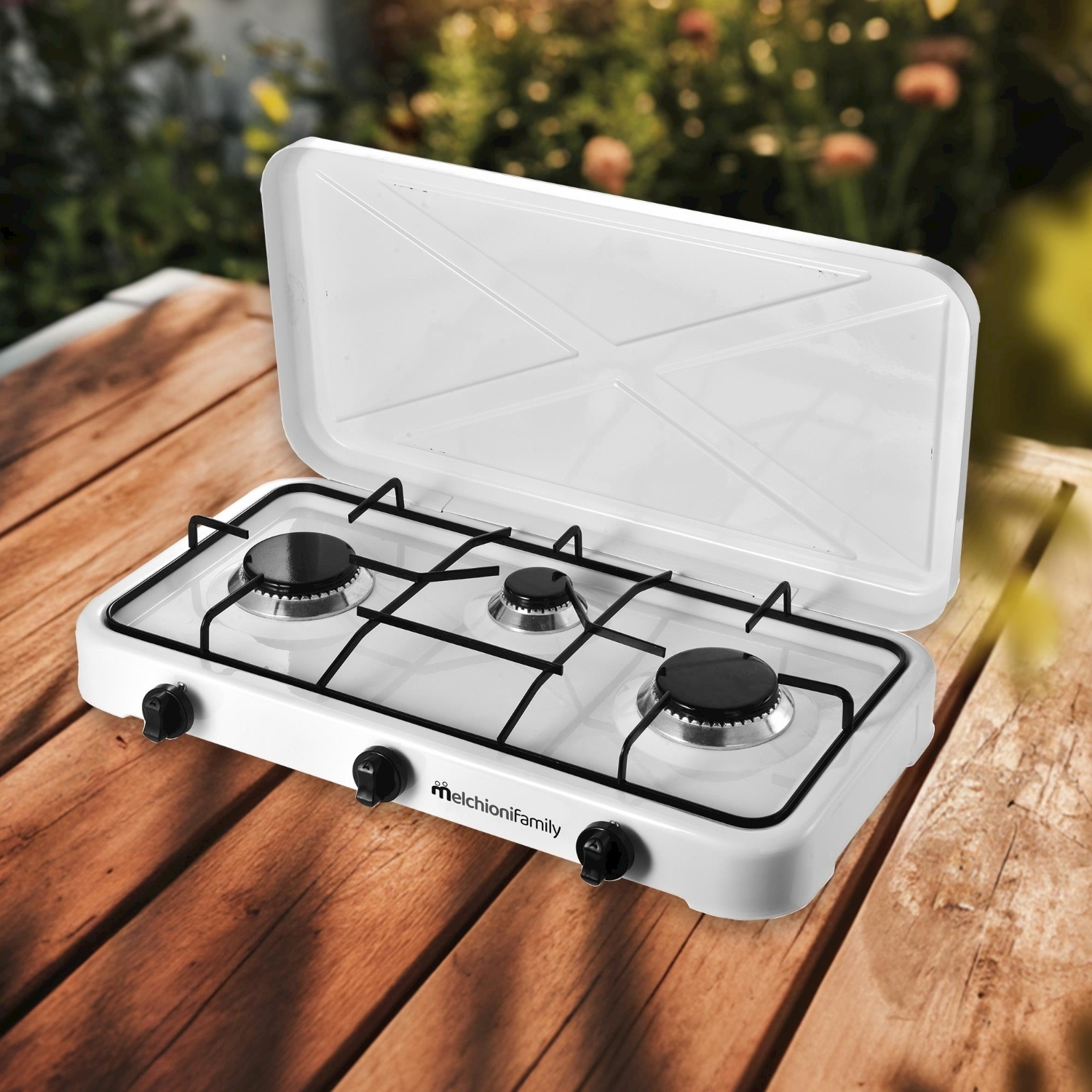 Fornello a gas portatile bianco su tavolo di legno, coperchio aperto. Sfondo con piante verdi e sole.