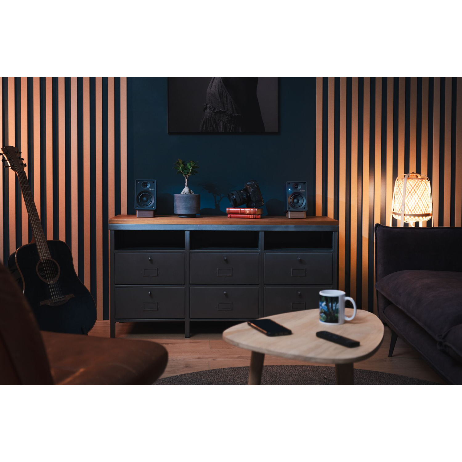 Woonkamerscène: Luidsprekers, gitaar en een kast met een lamp en een foto aan de muur.
