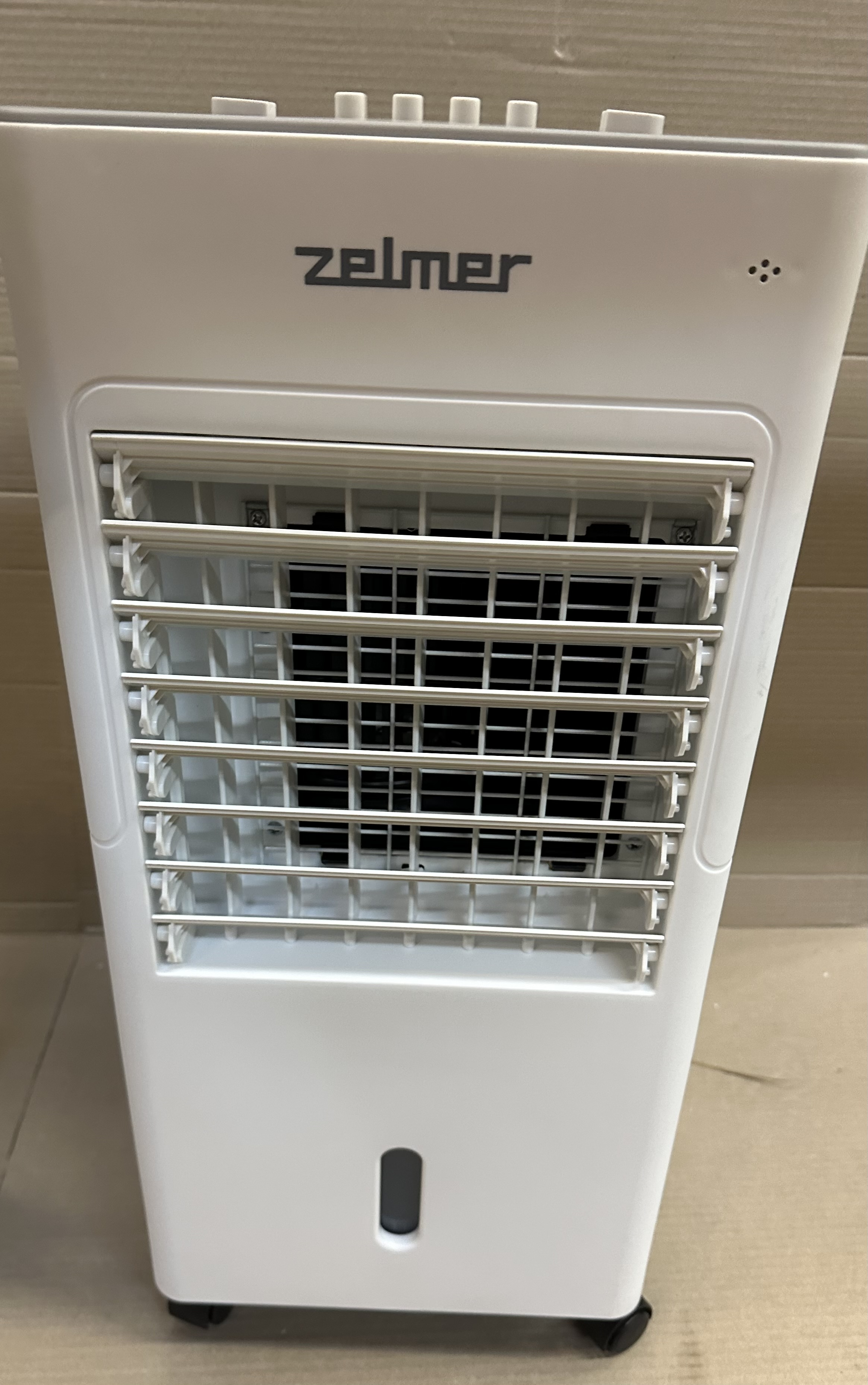 Biała chłodziarka powietrza z logo Zelmer. Posiada kratkę i kółka ułatwiające przemieszczanie.
