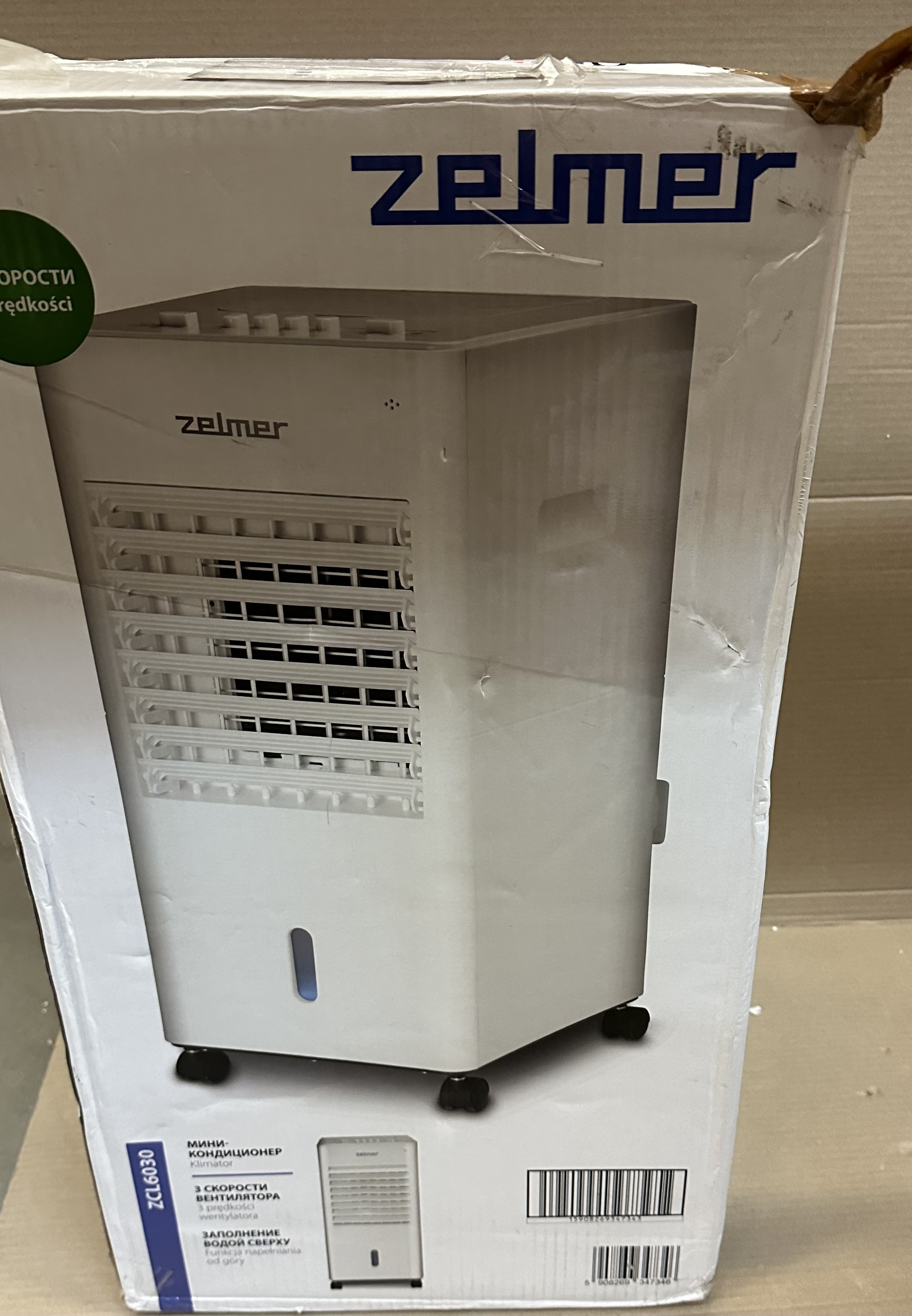 Biały klimatyzator w pudełku. Pudełko jest białe, z napisem 'zelmer' z przodu.