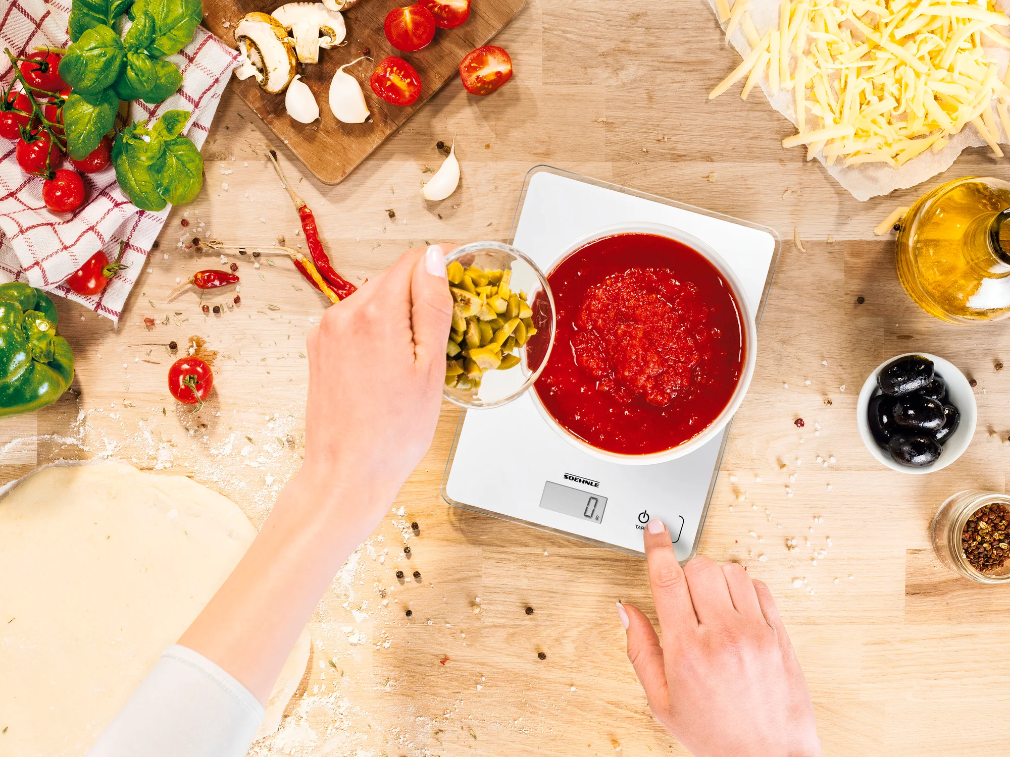Una persona aggiunge sottaceti a una ciotola di salsa di pomodoro. Ingredienti, bilancia e tagliere su tavolo.
