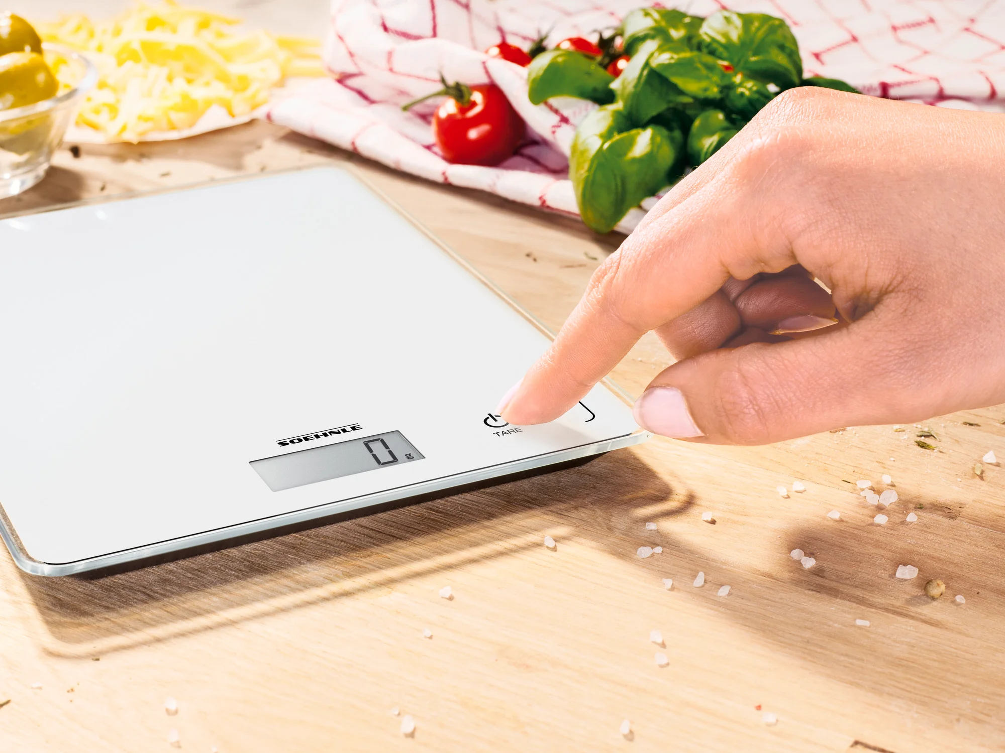 Mano che preme un pulsante su una bilancia digitale bianca, con cibo intorno.