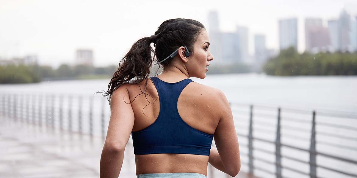 Kobieta biegnąca, w niebieskim staniku sportowym i słuchawkach, w pobliżu wody i panoramy miasta.