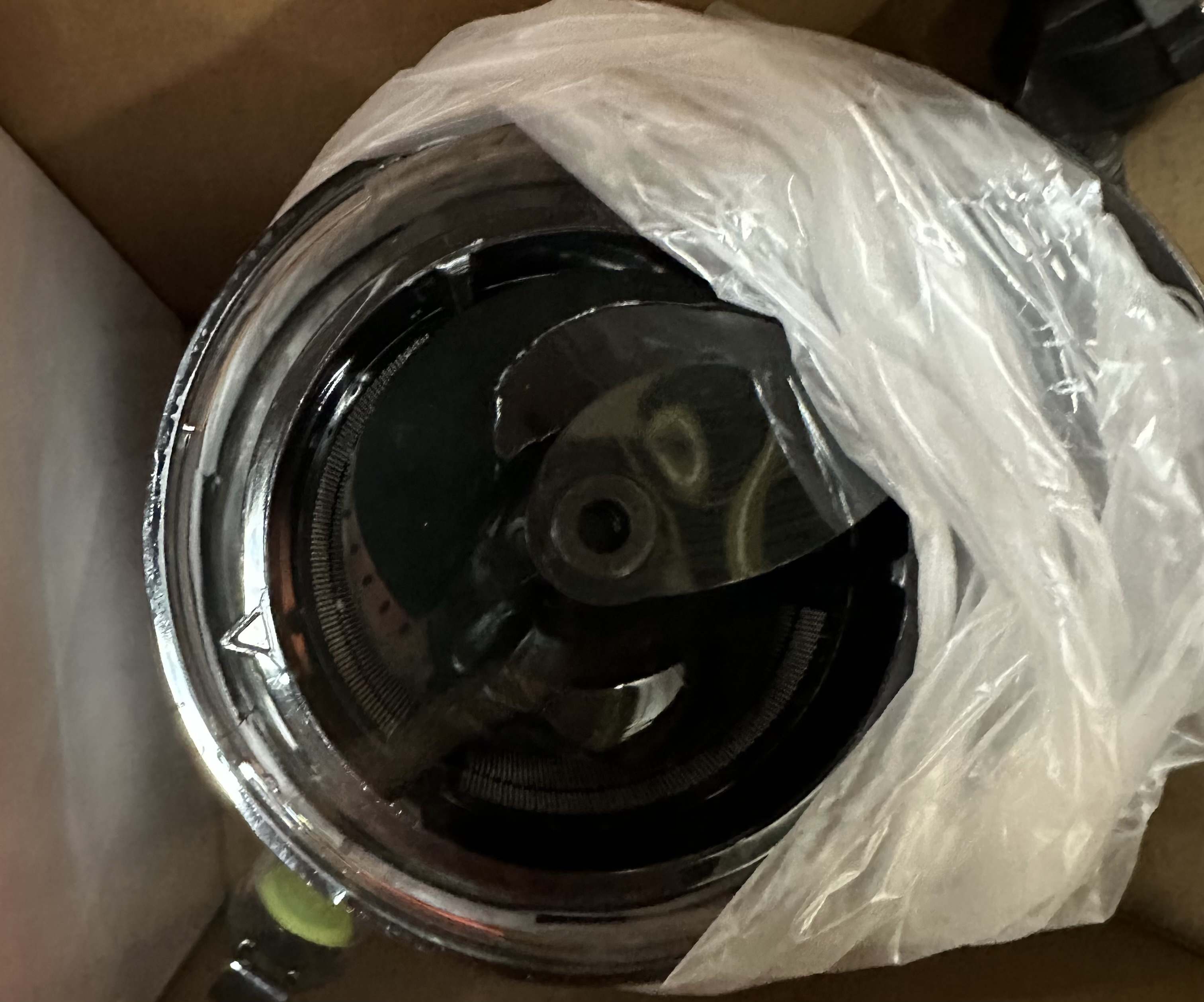 Zbliżenie błyszczącego, metalowego przedmiotu owiniętego w przezroczysty plastik, w kartonowym pudełku.