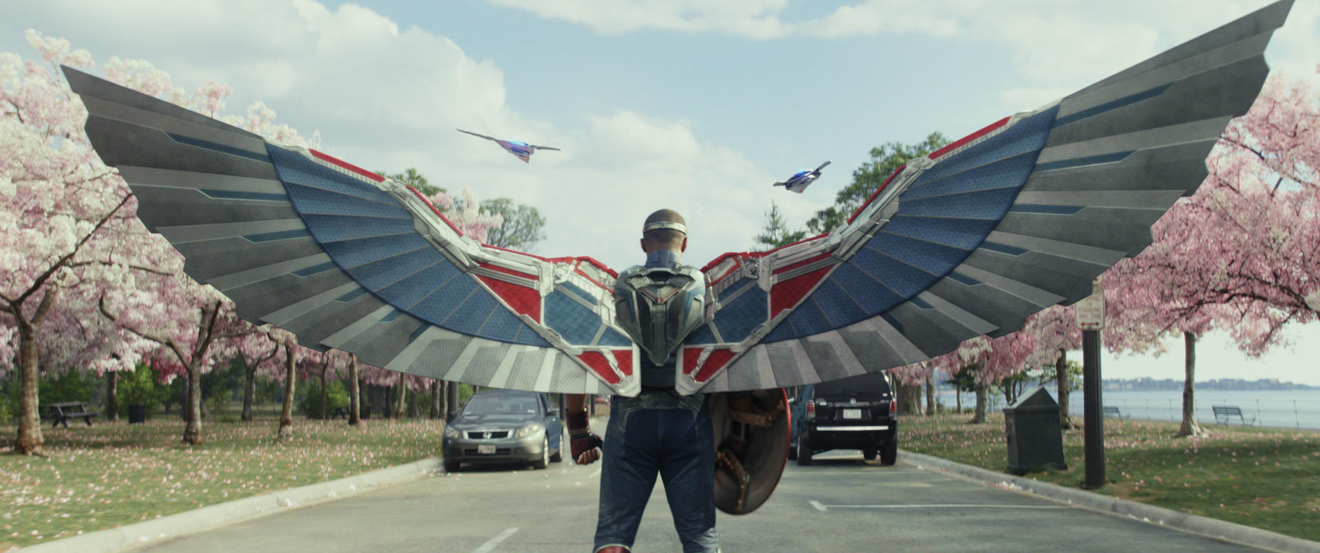 Eine Person in einem Captain America-Anzug mit Flügeln steht auf einer Straße mit Kirschblütenbäumen.