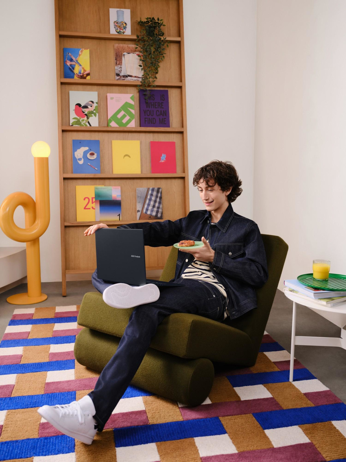 Man sitting on an olive green chair, working on laptop, with a snack and a lamp.