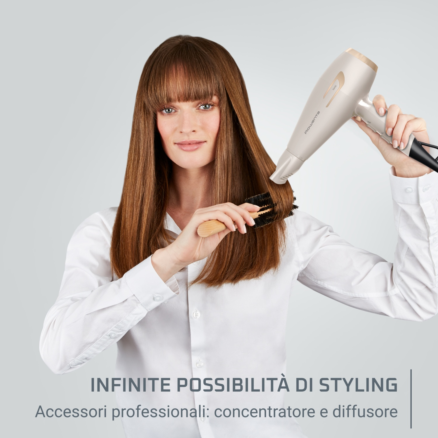 Donna che si acconcia i capelli con un asciugacapelli e una spazzola. Camicia bianca, sfondo chiaro.