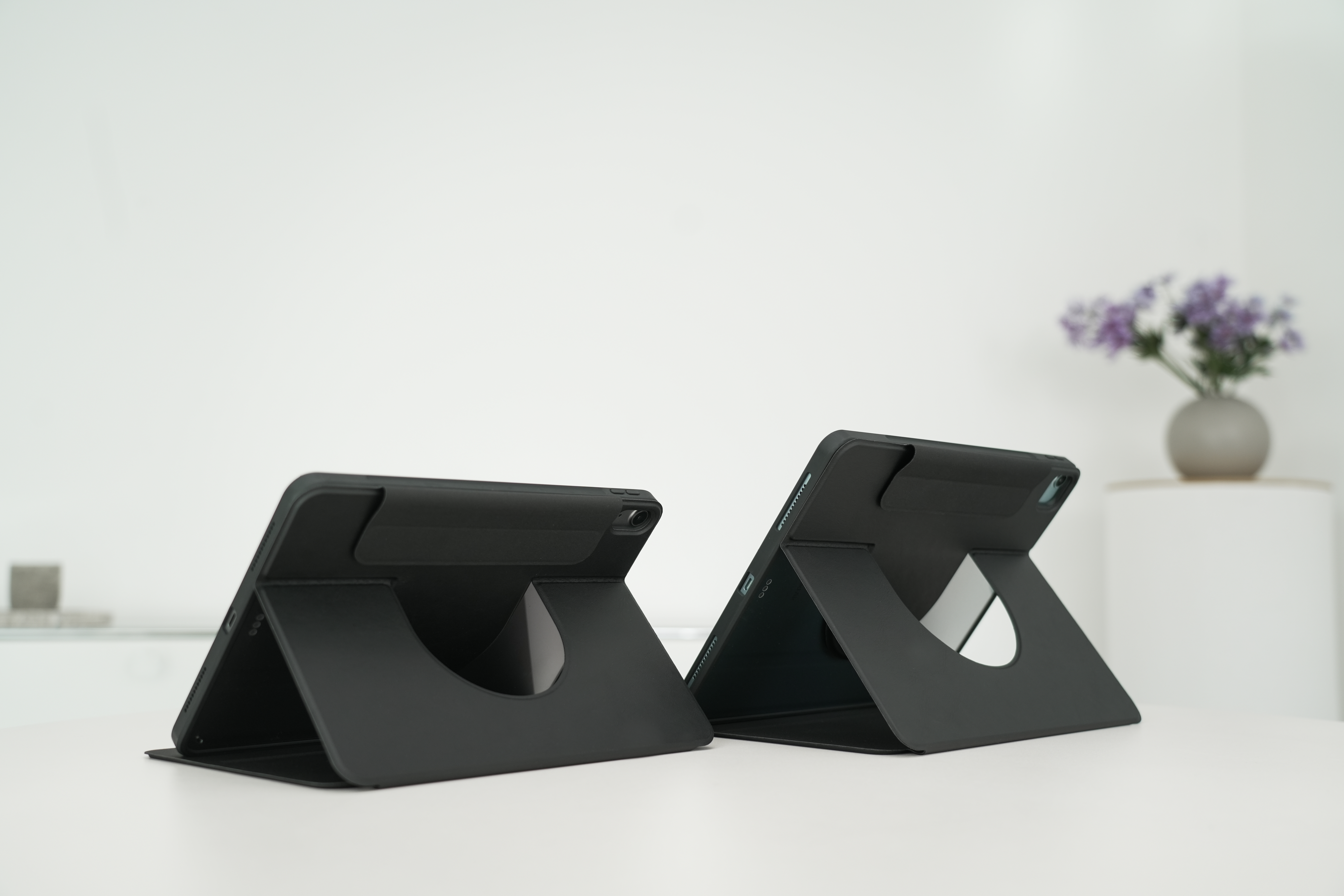 Zwei schwarze Tablet-Ständer auf einem weißen Tisch, mit weißer Wand und Blumen im Hintergrund.