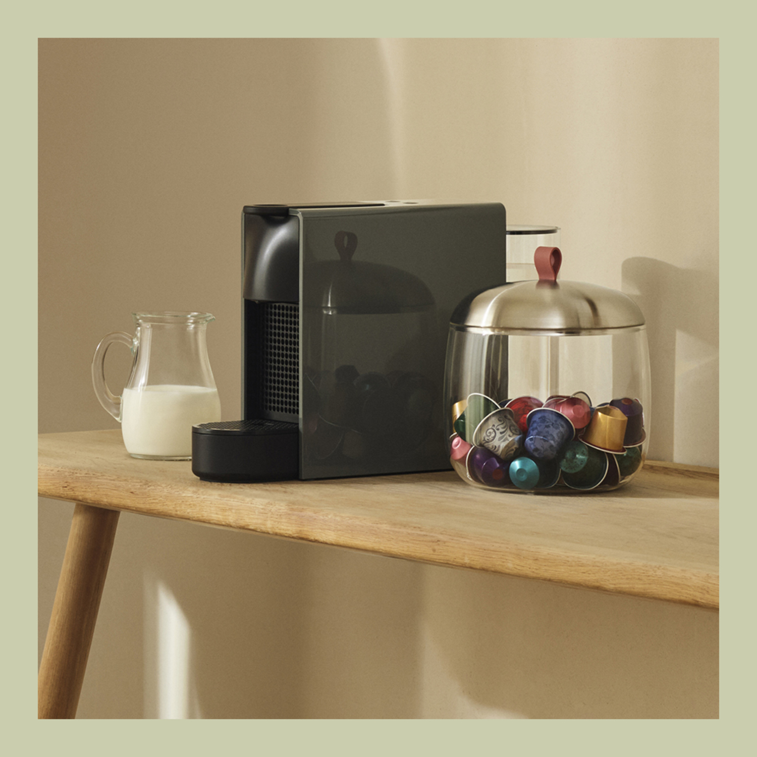 Coffee machine, milk jug, glass container with capsules. Wooden surface. Neutral colors. Warm tones.