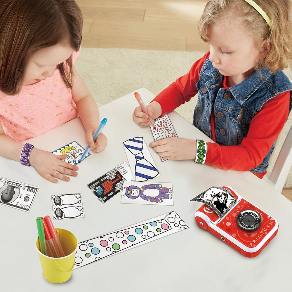 Due bambini che disegnano su carta con pennarelli e una fotocamera Printcam rossa.