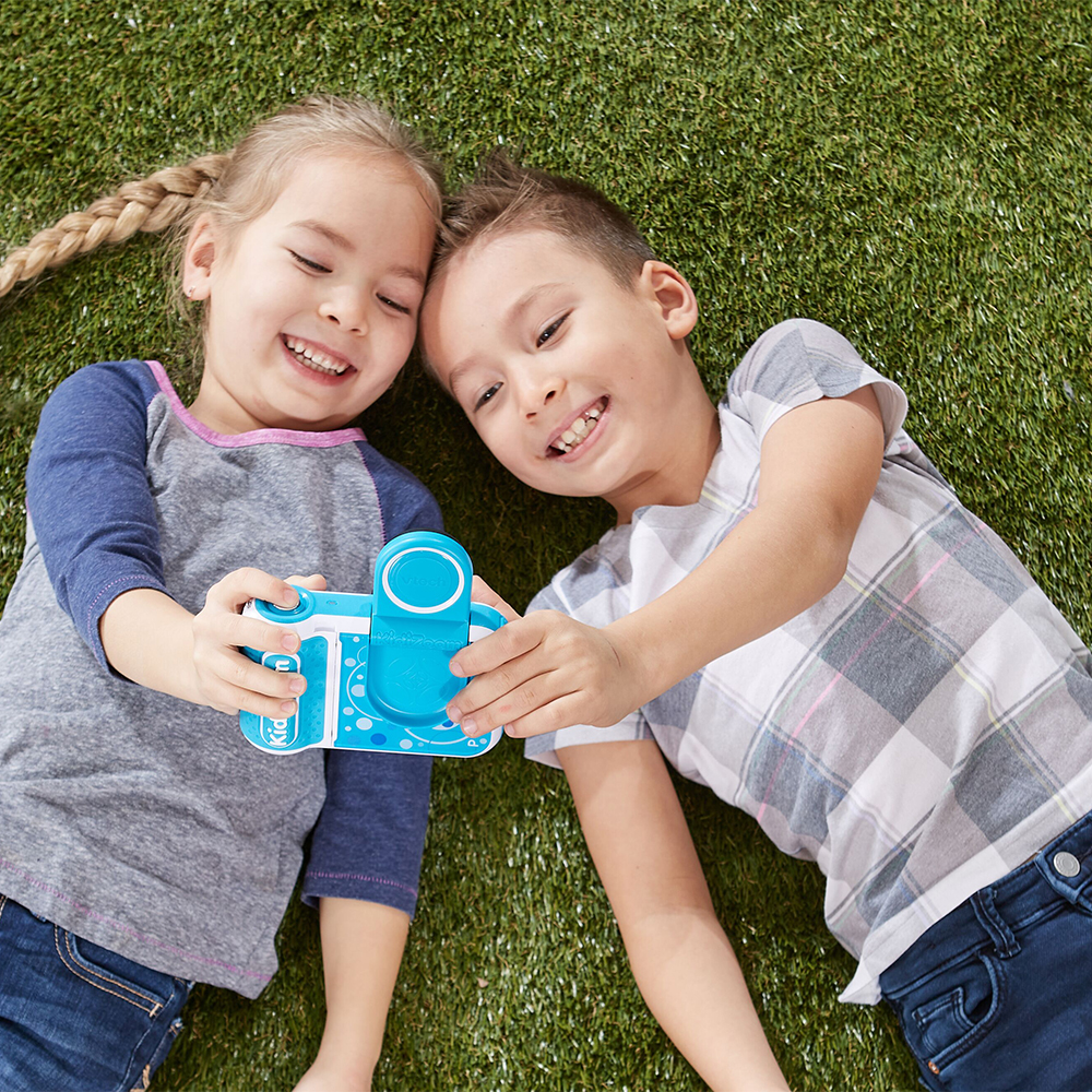 Zwei Kinder auf Gras, halten eine blaue Spielzeugkamera und lächeln.