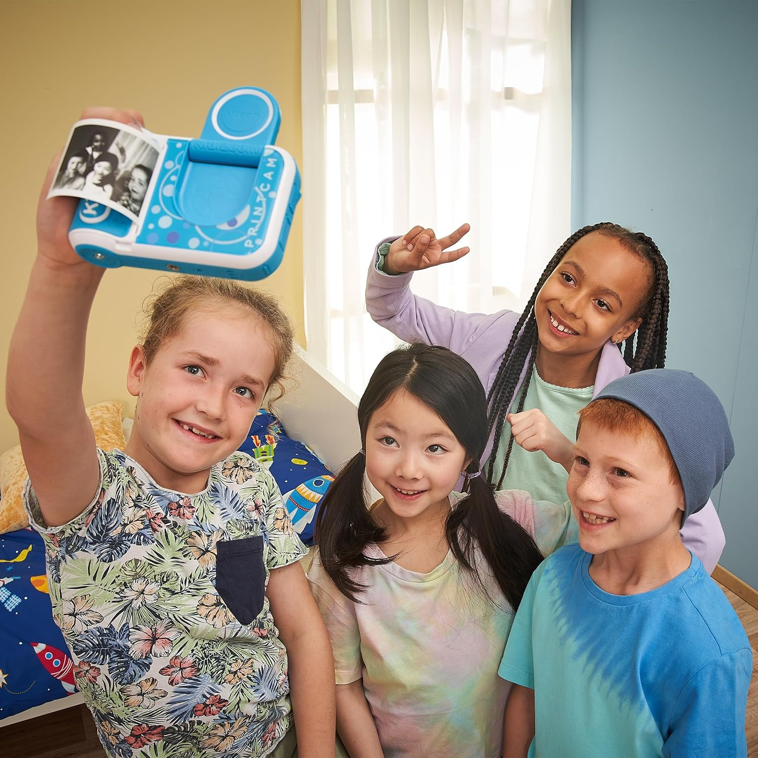 Vier Kinder in einem Schlafzimmer, eines hält eine Druckkamera, andere lächeln und posieren.