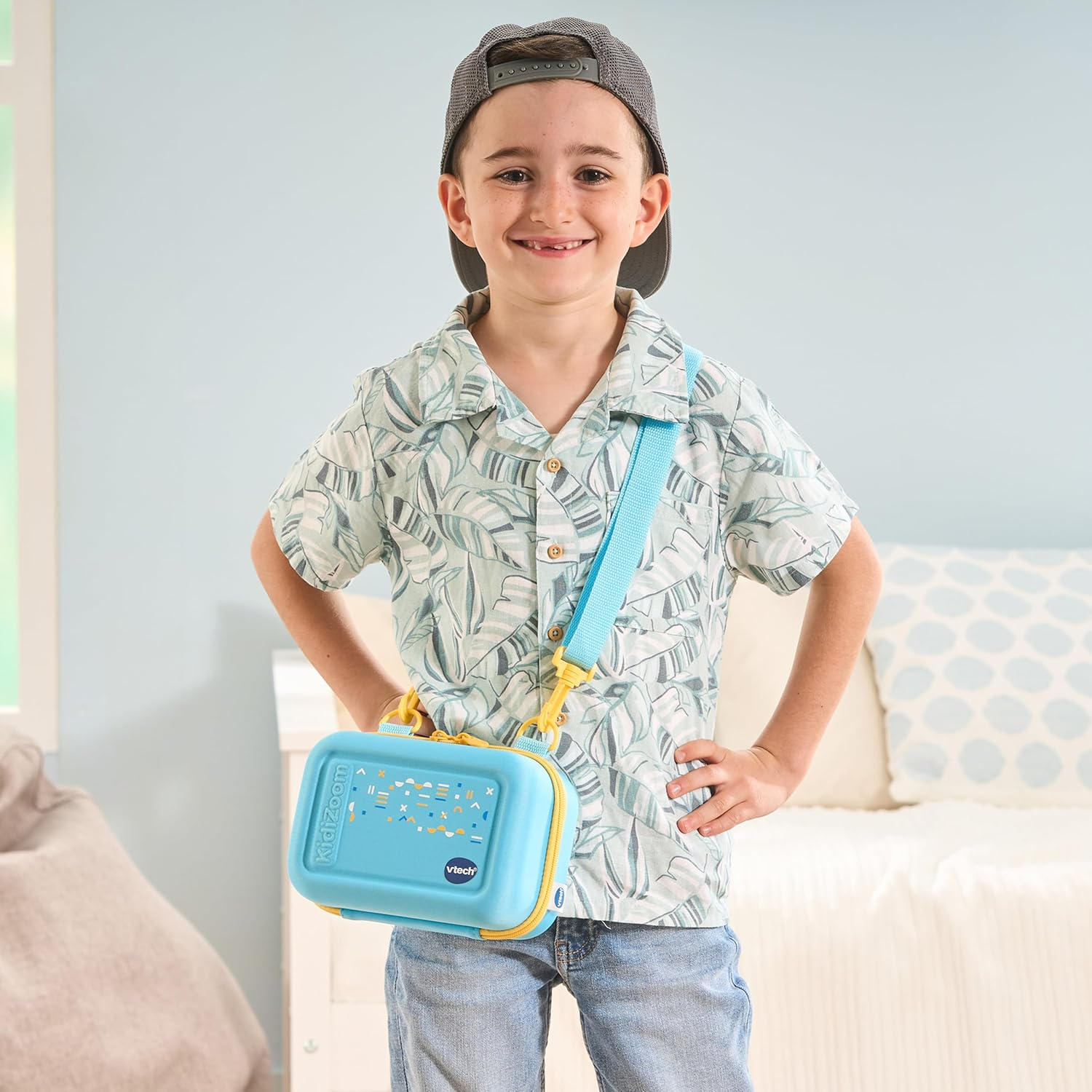 Un garçon souriant portant une casquette grise et un sac à jouets bleu avec une sangle jaune. Il est à l'intérieur.