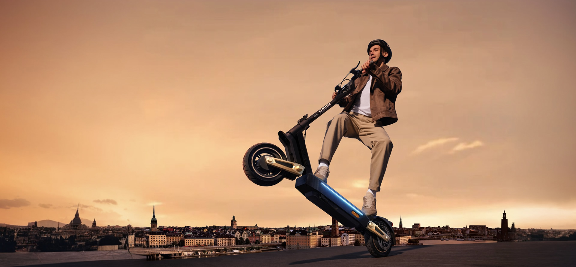 Mężczyzna na hulajnodze elektrycznej, pochylony do tyłu. Tło to krajobraz miejski z zachodem słońca.