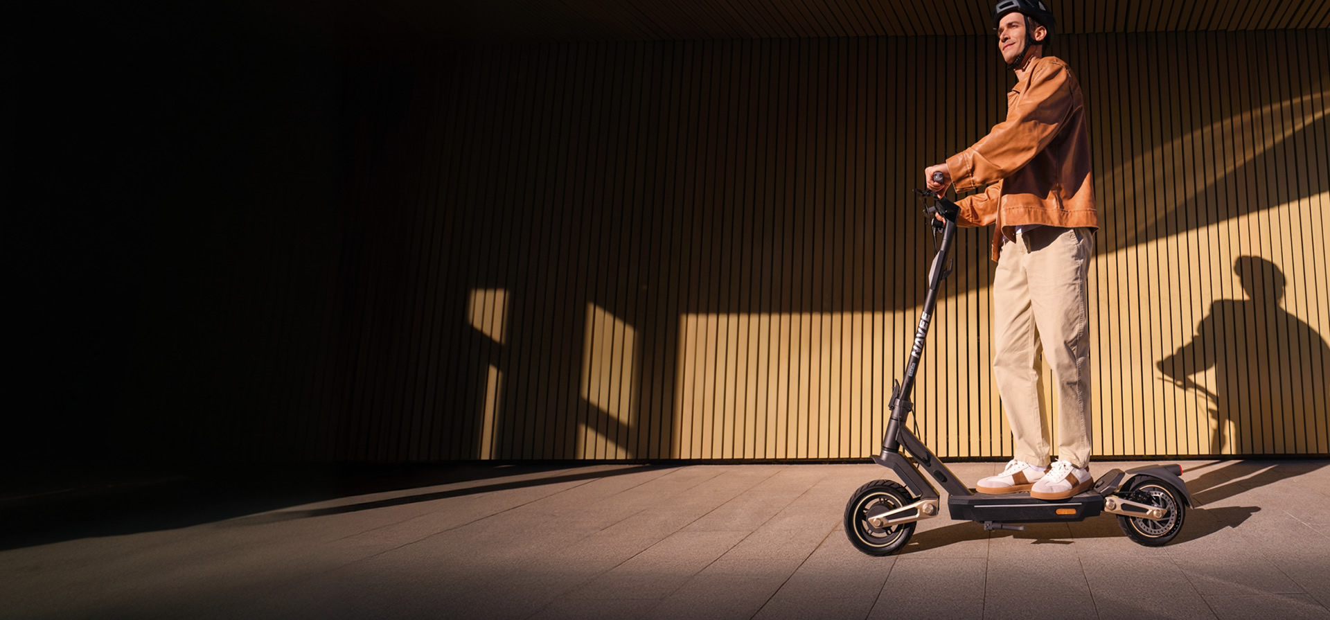 Mężczyzna na czarno-złotej hulajnodze, w kasku, na zewnątrz.