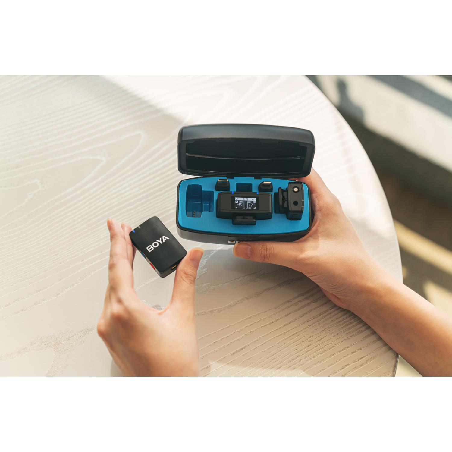 Handen die een zwart audioapparaat met accessoires vasthouden. Het apparaat zit in een draagtas.