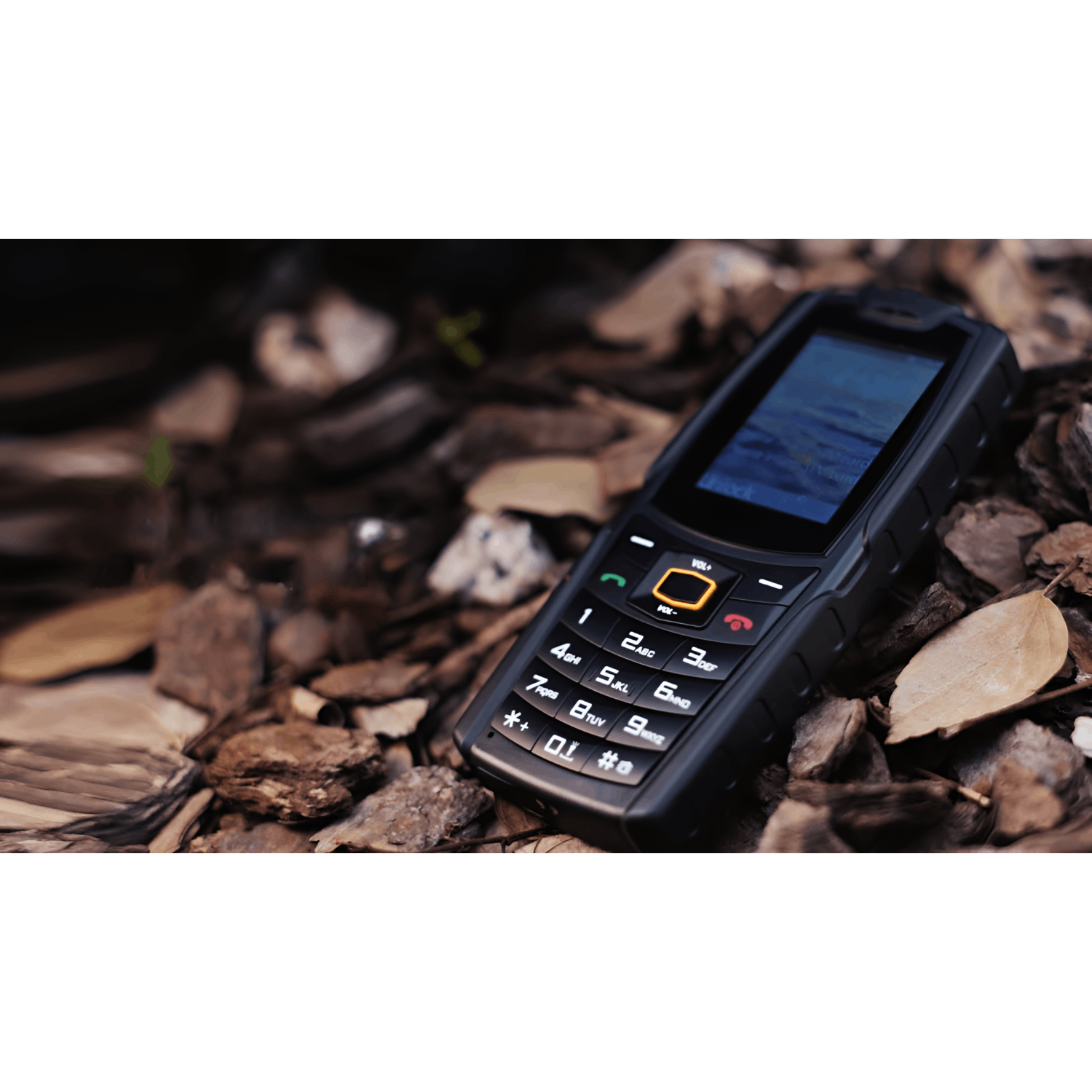 Un téléphone portable noir et robuste est posé sur des copeaux de bois. L'écran affiche une scène d'eau bleue. Des boutons et des chiffres sont visibles.