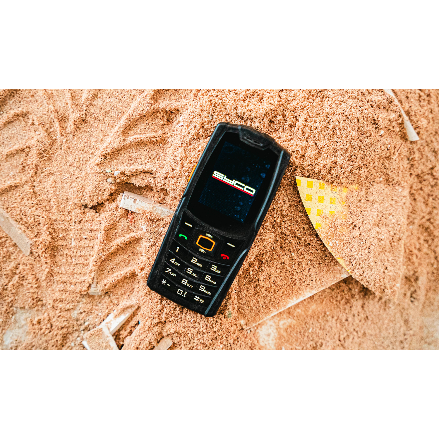 Un téléphone SYCO noir repose sur de la sciure de bois. Un objet jaune et blanc est partiellement visible. Des empreintes dans la sciure.