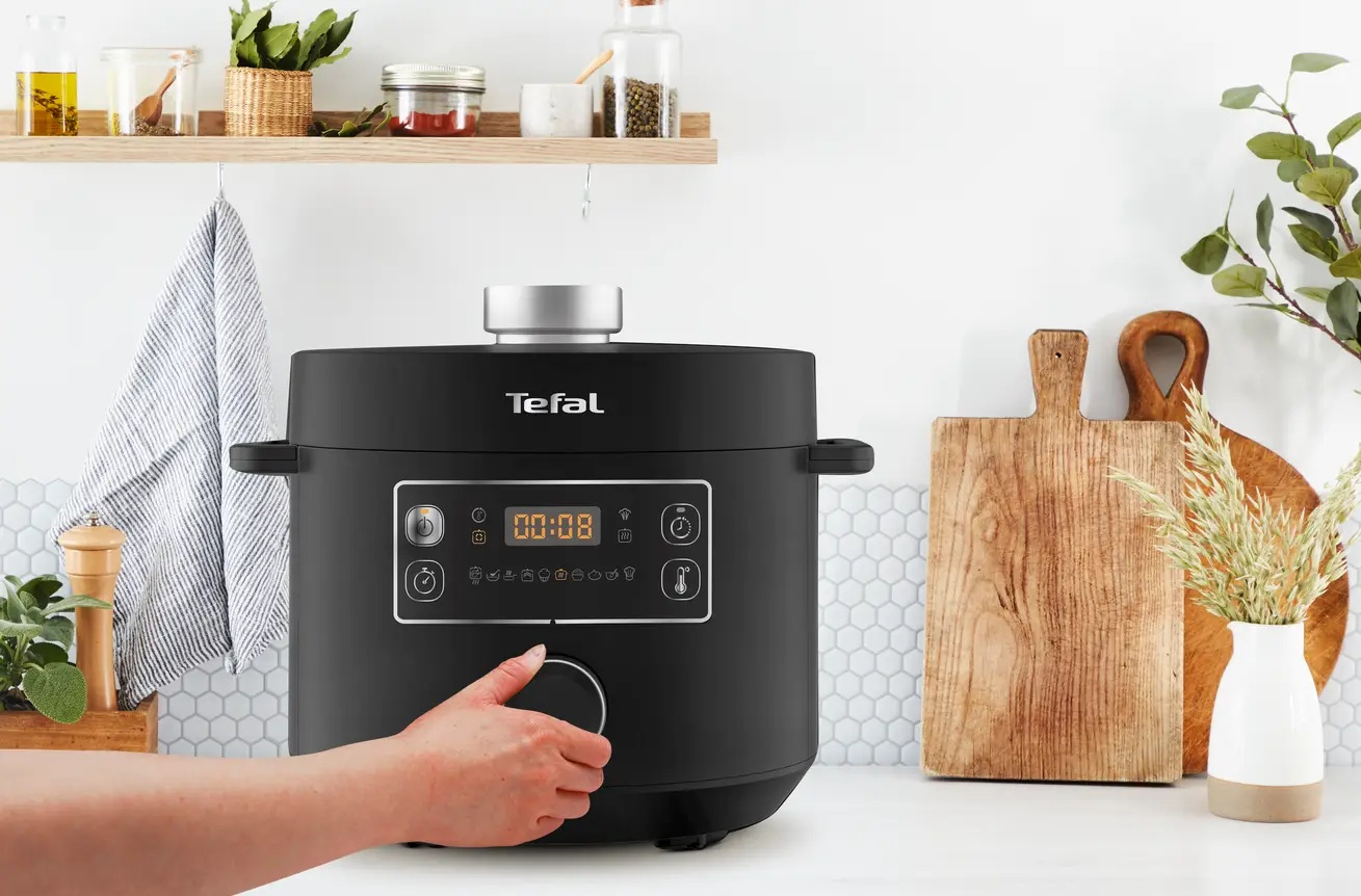 Czarny multicooker Tefal z wyświetlaczem cyfrowym. Ręka obraca pokrętło. Tło kuchenne z półkami.