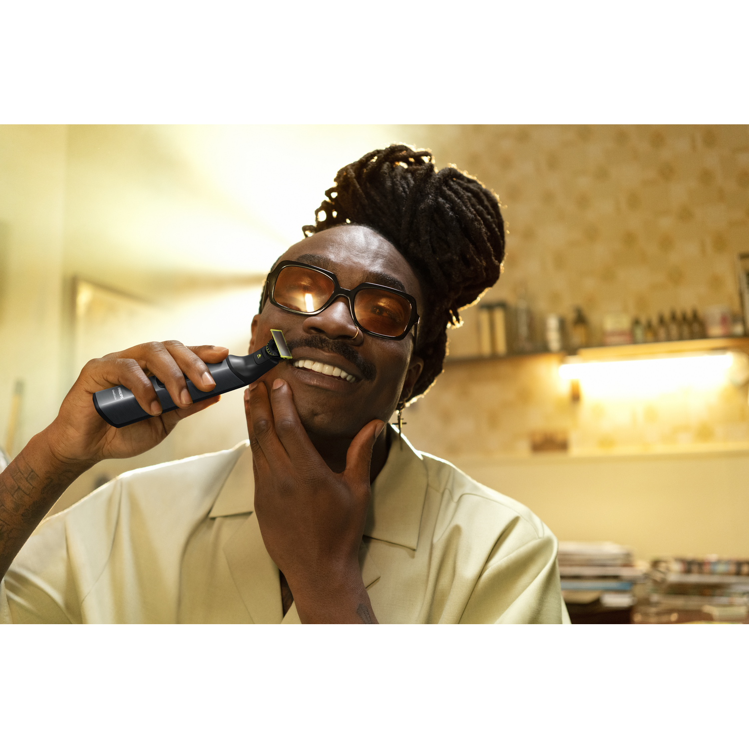 A man with dreadlocks is shaving his neck with a black trimmer, smiling.