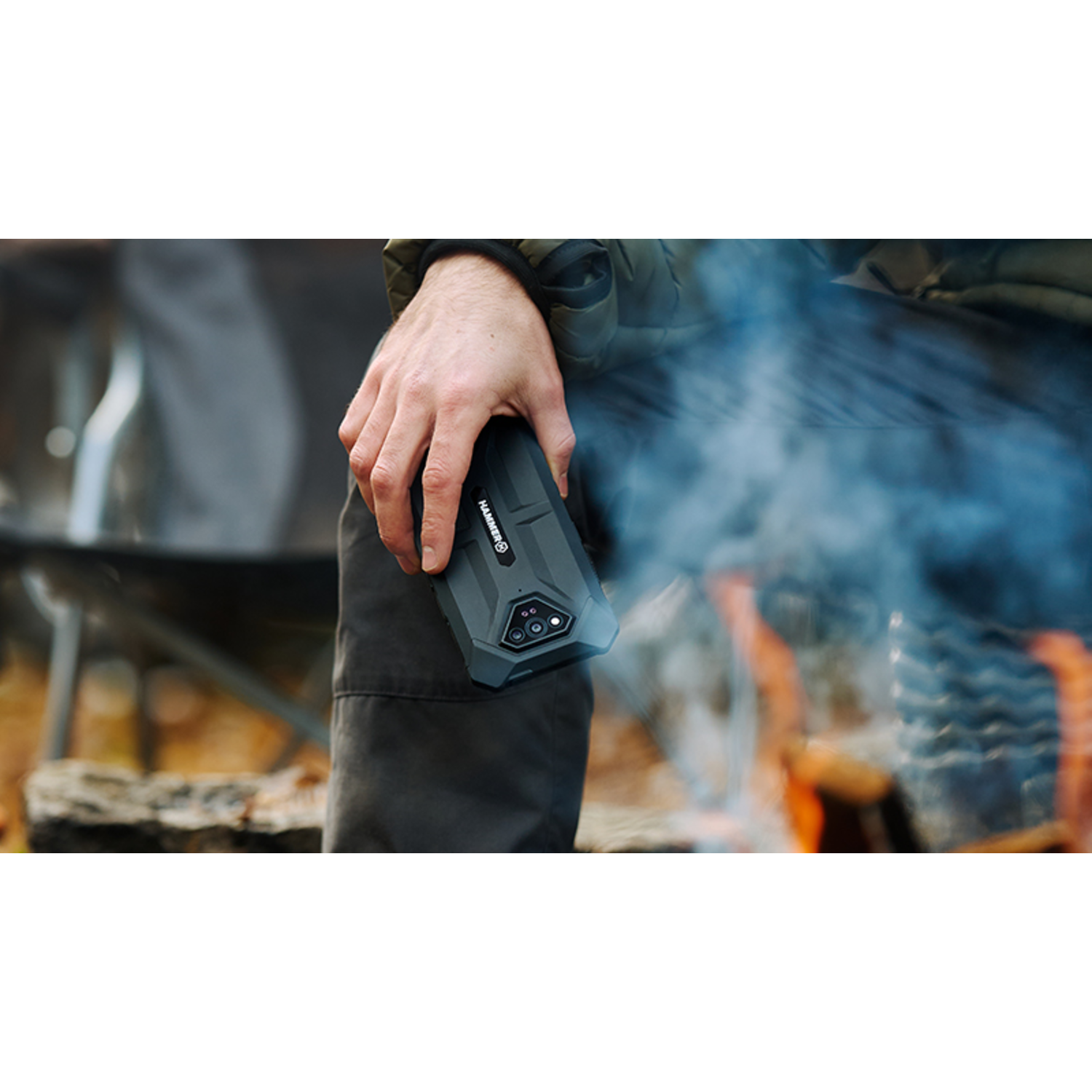 Een persoon houdt een zwarte smartphone vast in de buurt van een kampvuur. Er komt rook op. Op de telefoon staat het woord HAMMER.