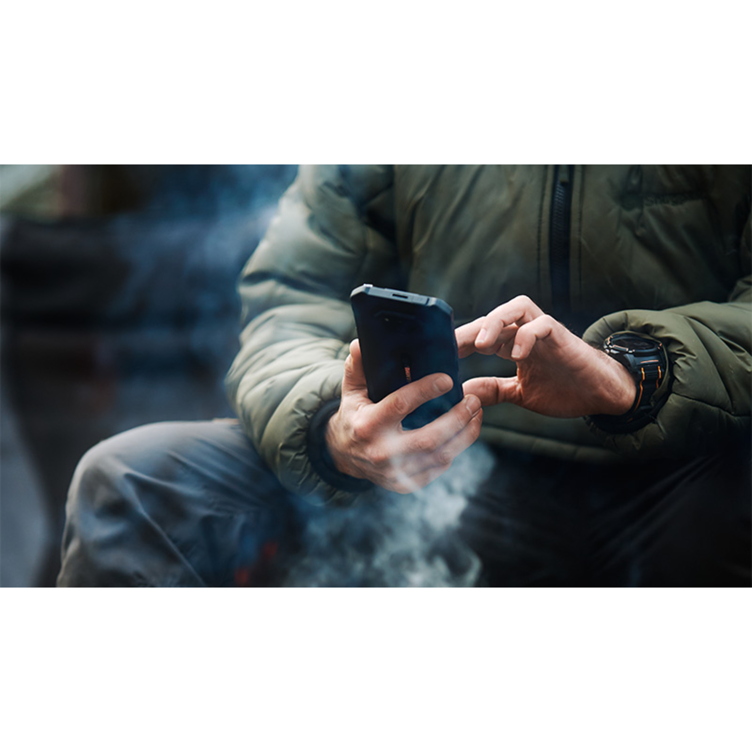 A person wearing a green jacket and watch is using a black smartphone. Smoke is visible in the background.