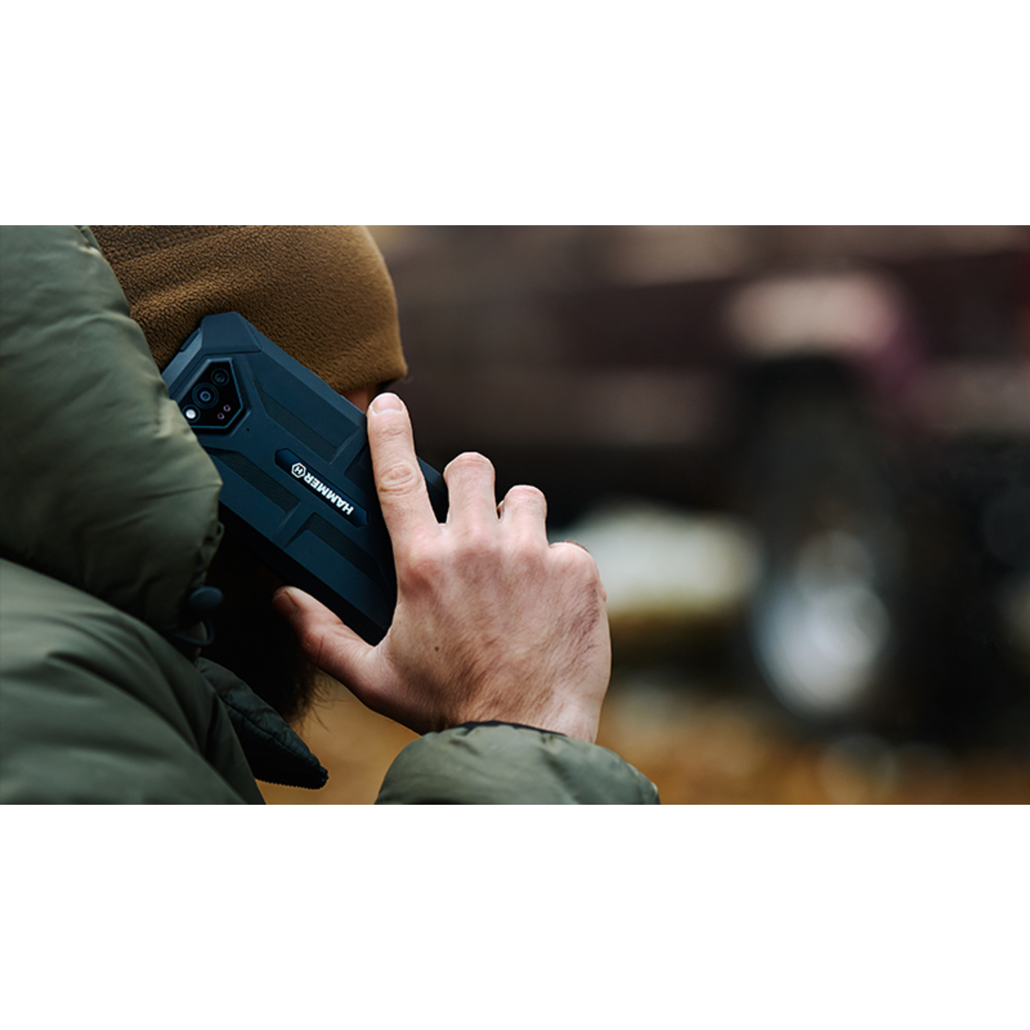 A person wearing a green jacket and brown beanie holds a black smartphone to their ear. Blurred background with a vehicle.