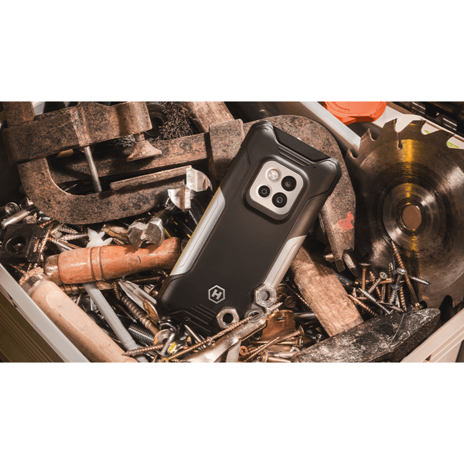 Black smartphone with multiple camera lenses lies in a toolbox. Tools like saw blades, screws, and clamps surround it.