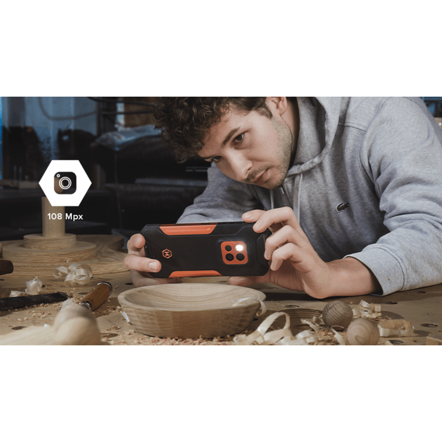 Een man in een grijze hoodie bekijkt een zwart-oranje telefoon. Houtbewerkingsgereedschap, houtkrullen en een houten kom liggen op tafel.