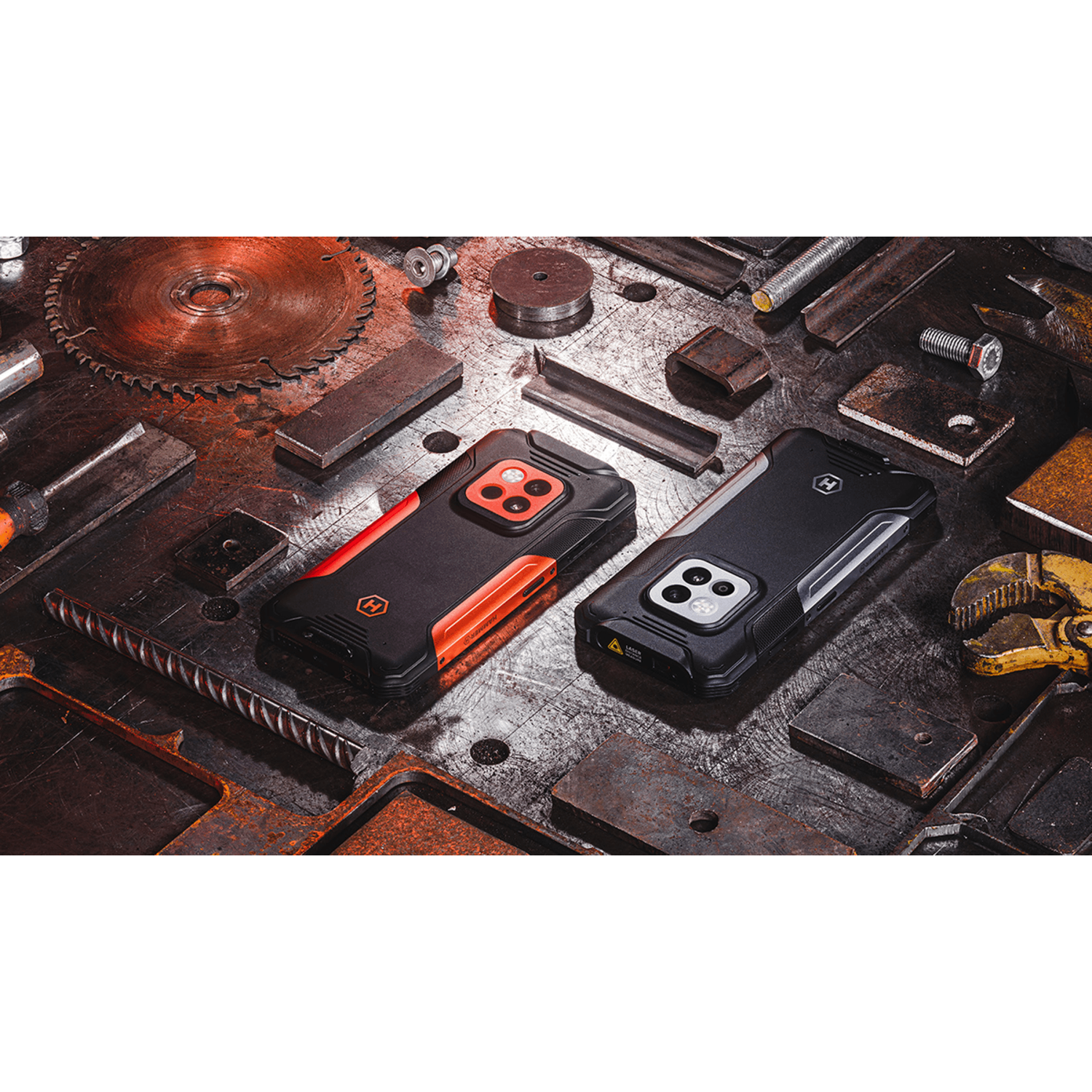Twee zwarte en oranje robuuste smartphones liggen op een metalen oppervlak met gereedschap en metalen componenten verspreid.
