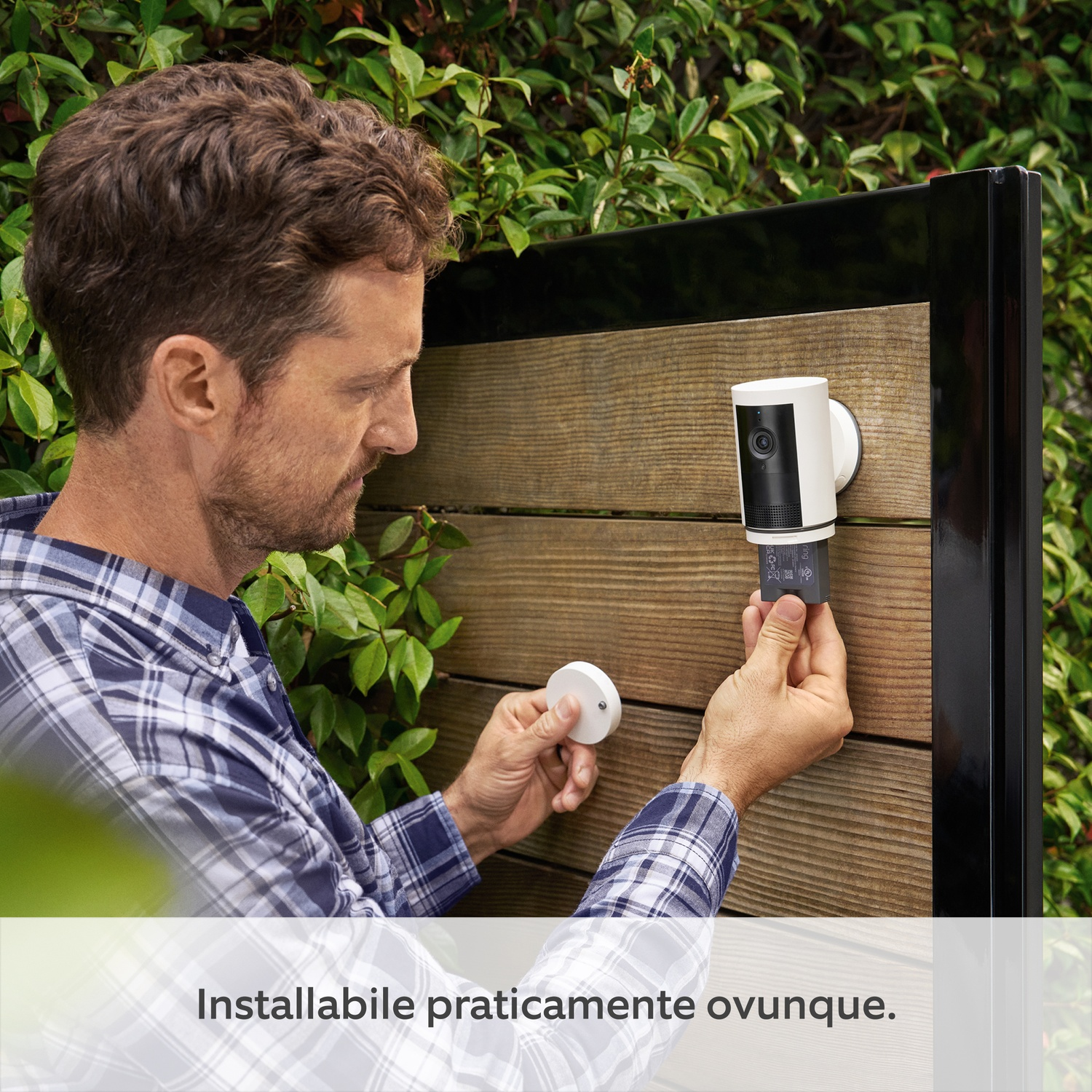 Un uomo che installa una telecamera di sicurezza su una recinzione di legno. La telecamera è bianca e nera.