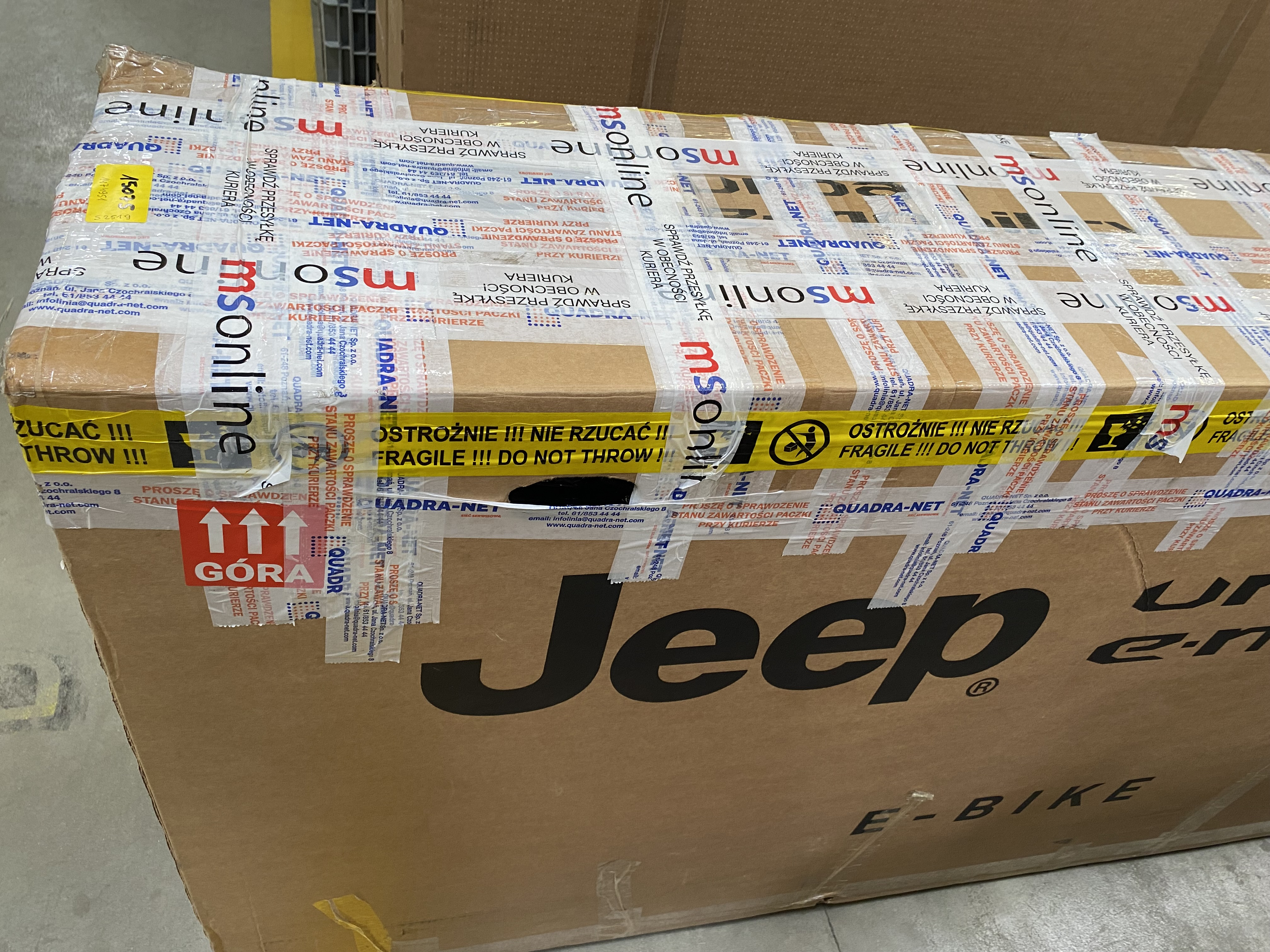 Kartonowe pudełko z taśmą i etykietami. Napis 'Jeep E-BIKE' i strzałki wskazujące górę.