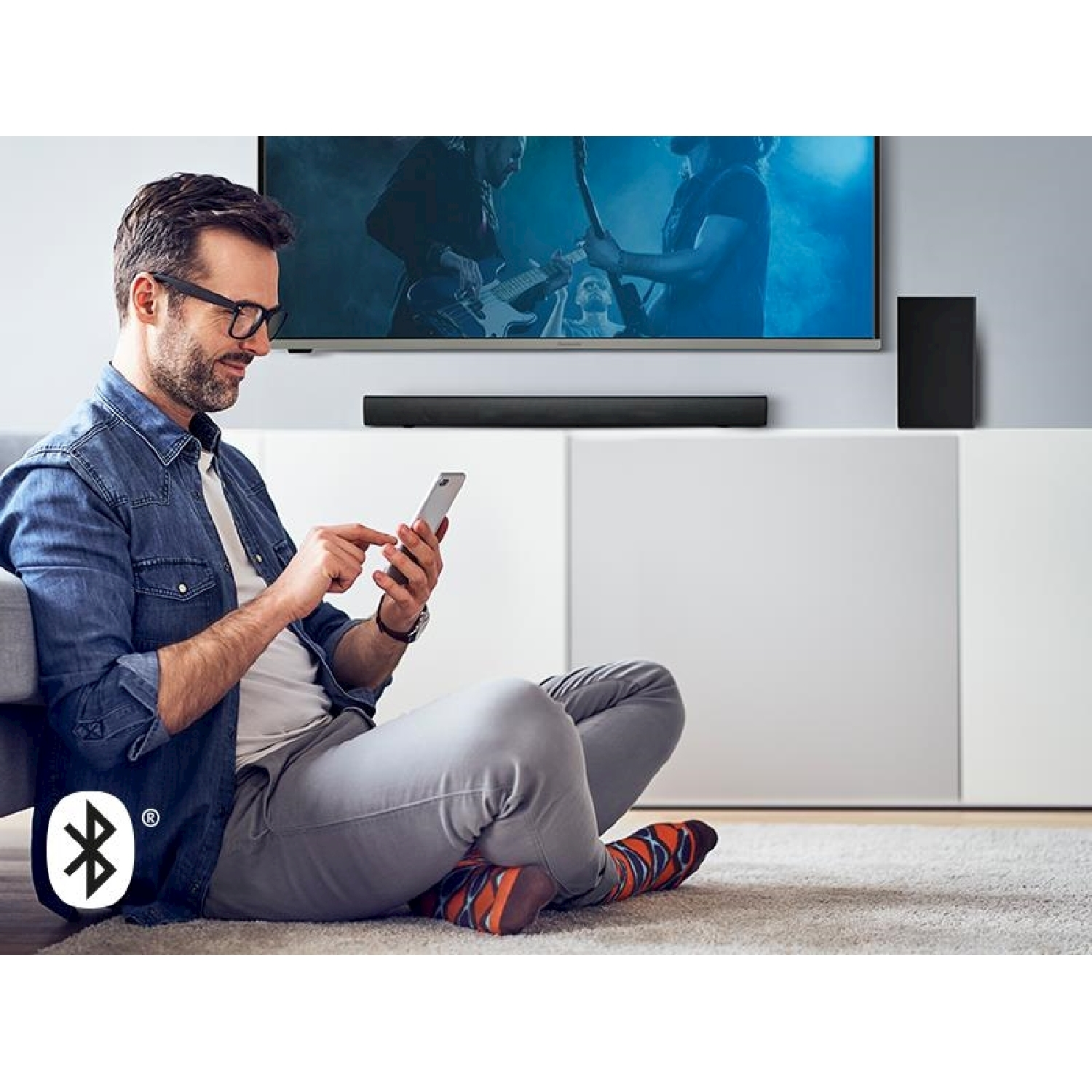 Un uomo è seduto, usa un telefono, di fronte a una TV con una soundbar.