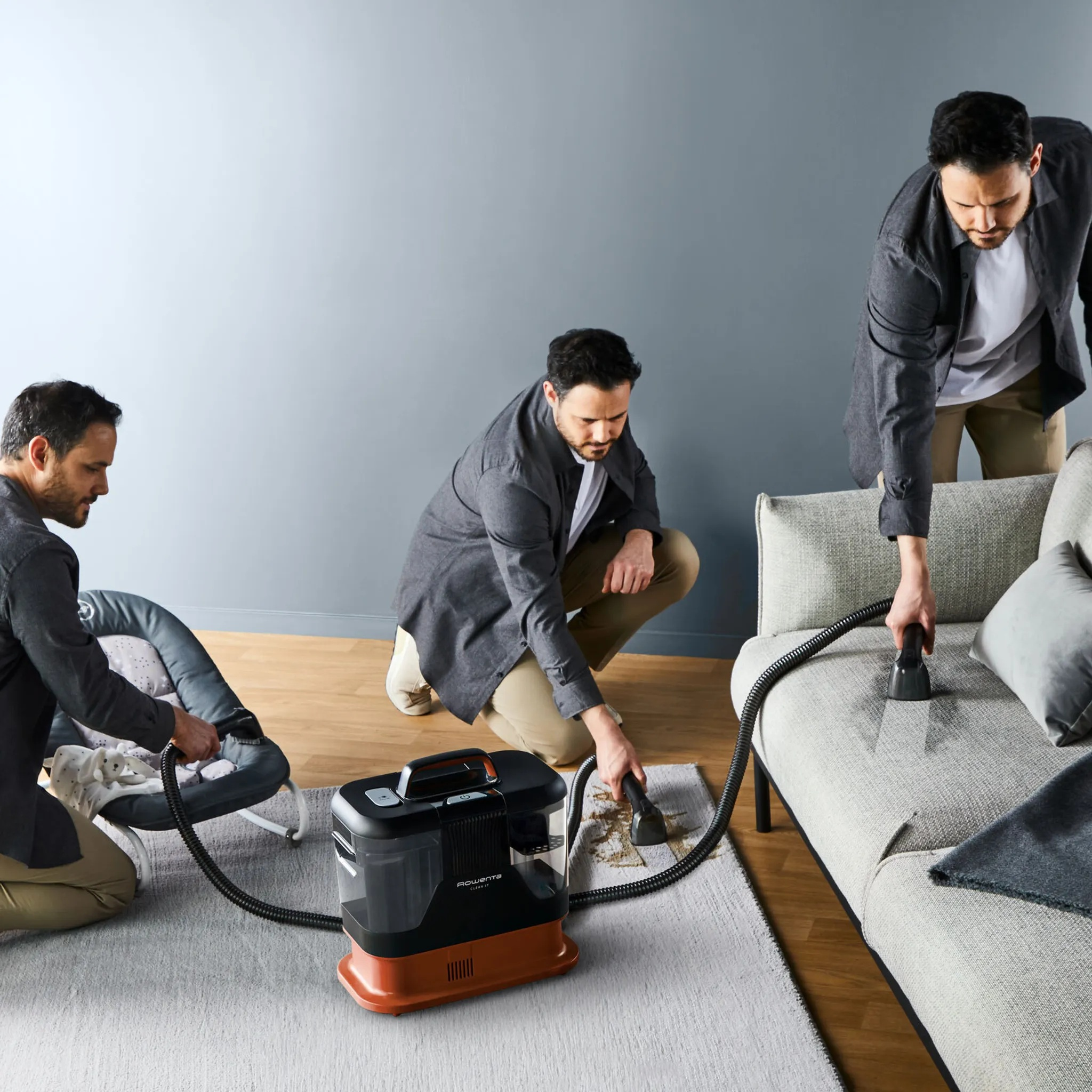 Trzech mężczyzn czyszczących sofę maszyną czyszczącą, szare tło.