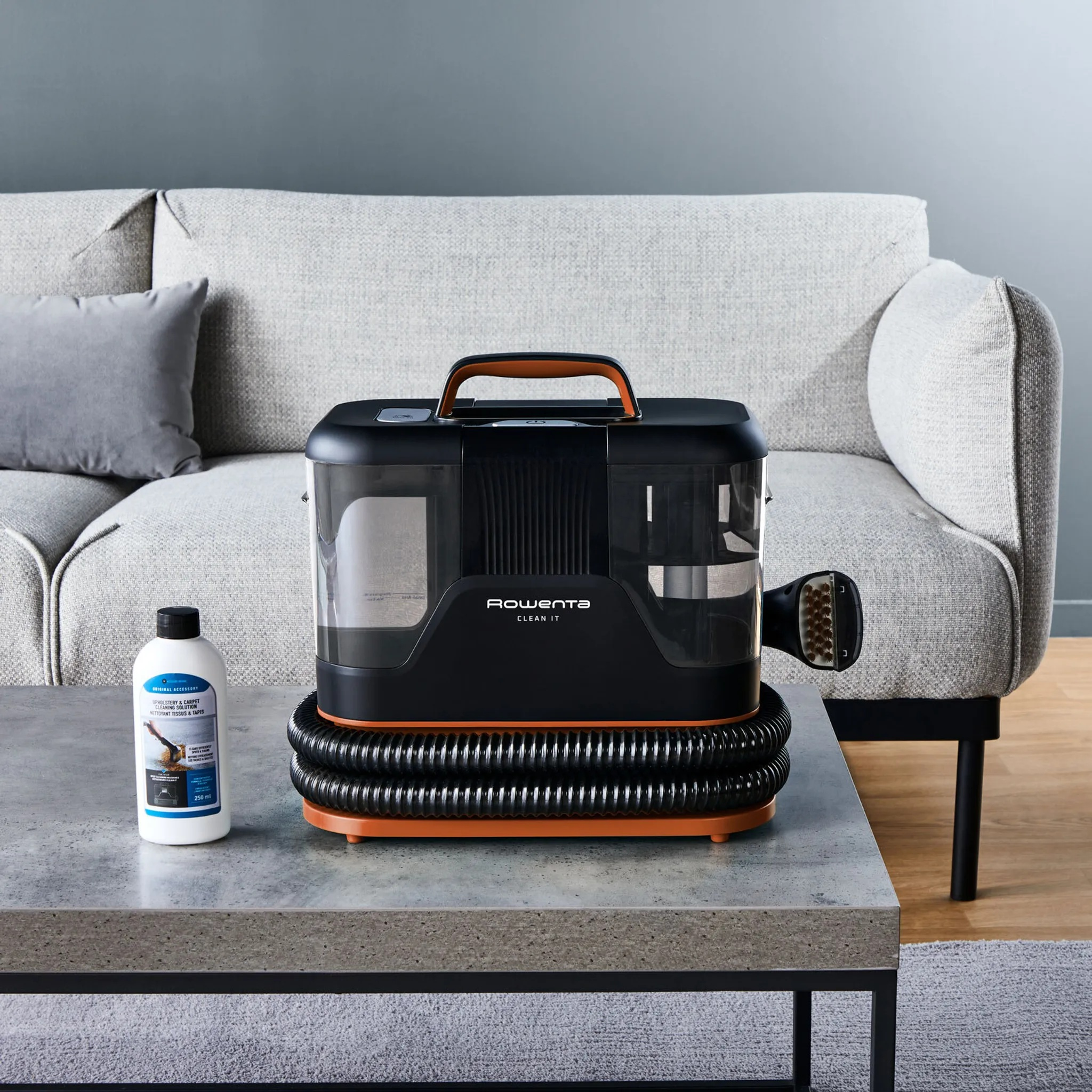 Czarny i pomarańczowy odkurzacz Rowenta na stole, z butelką środka czyszczącego i sofą w tle.