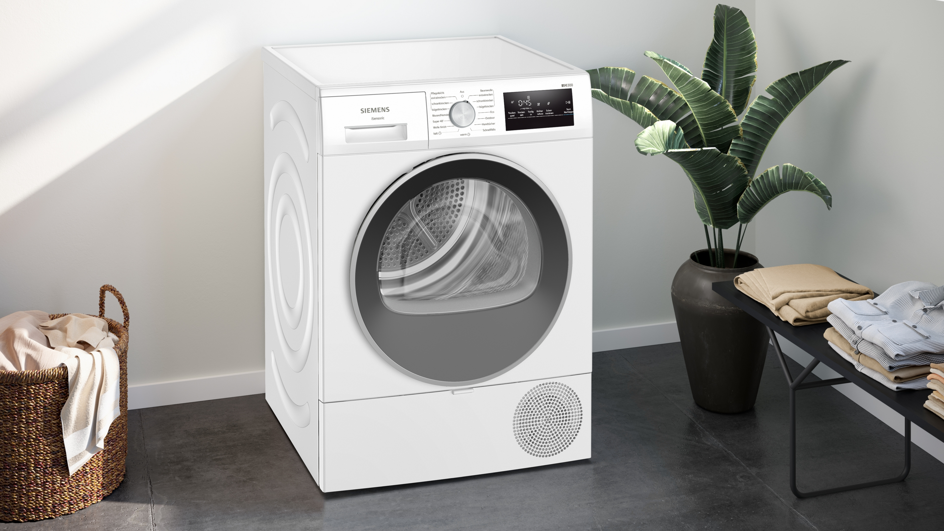 Ein weißer Siemens-Trockner mit schwarzer Tür, neben einer Pflanze und einem Tisch mit gefalteter Kleidung. Links ein Korb.