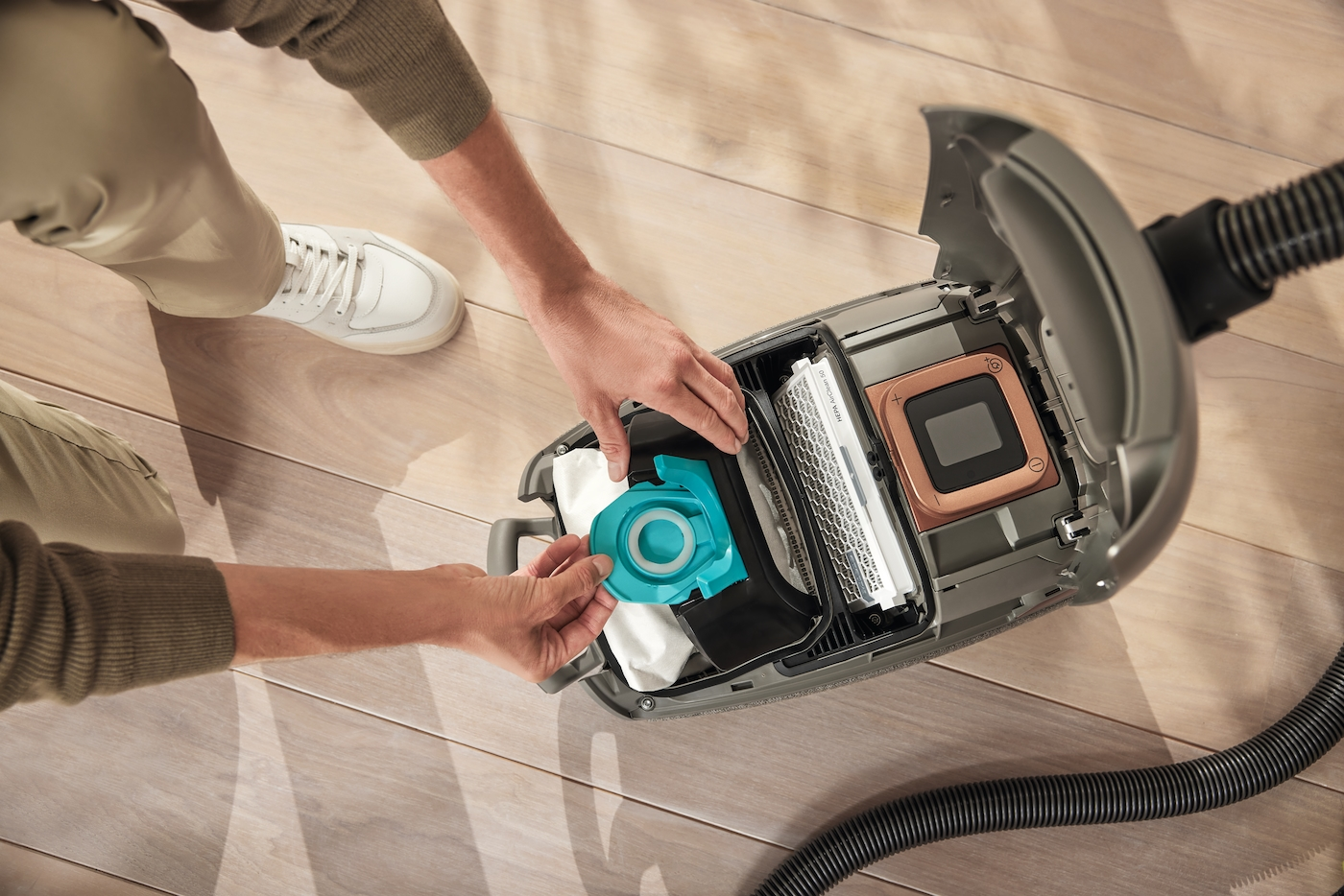 Eine Person ersetzt einen blauen Filter in einem offenen, grauen Staubsauger. Der Staubsauger steht auf einem Holzboden, ein Schuh ist sichtbar.