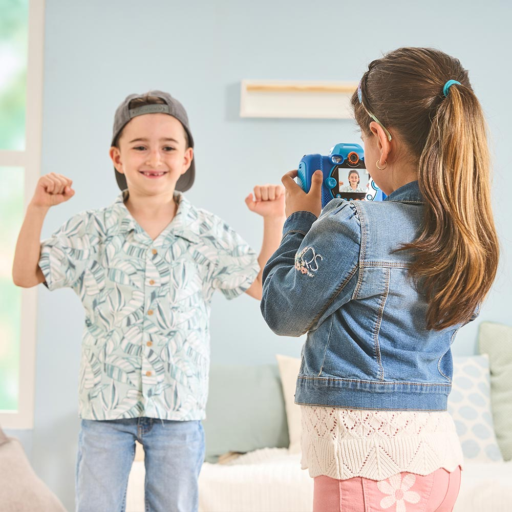 Zwei Kinder, eines macht ein Foto mit einer blauen Kamera, das andere spannt die Muskeln an und lächelt.